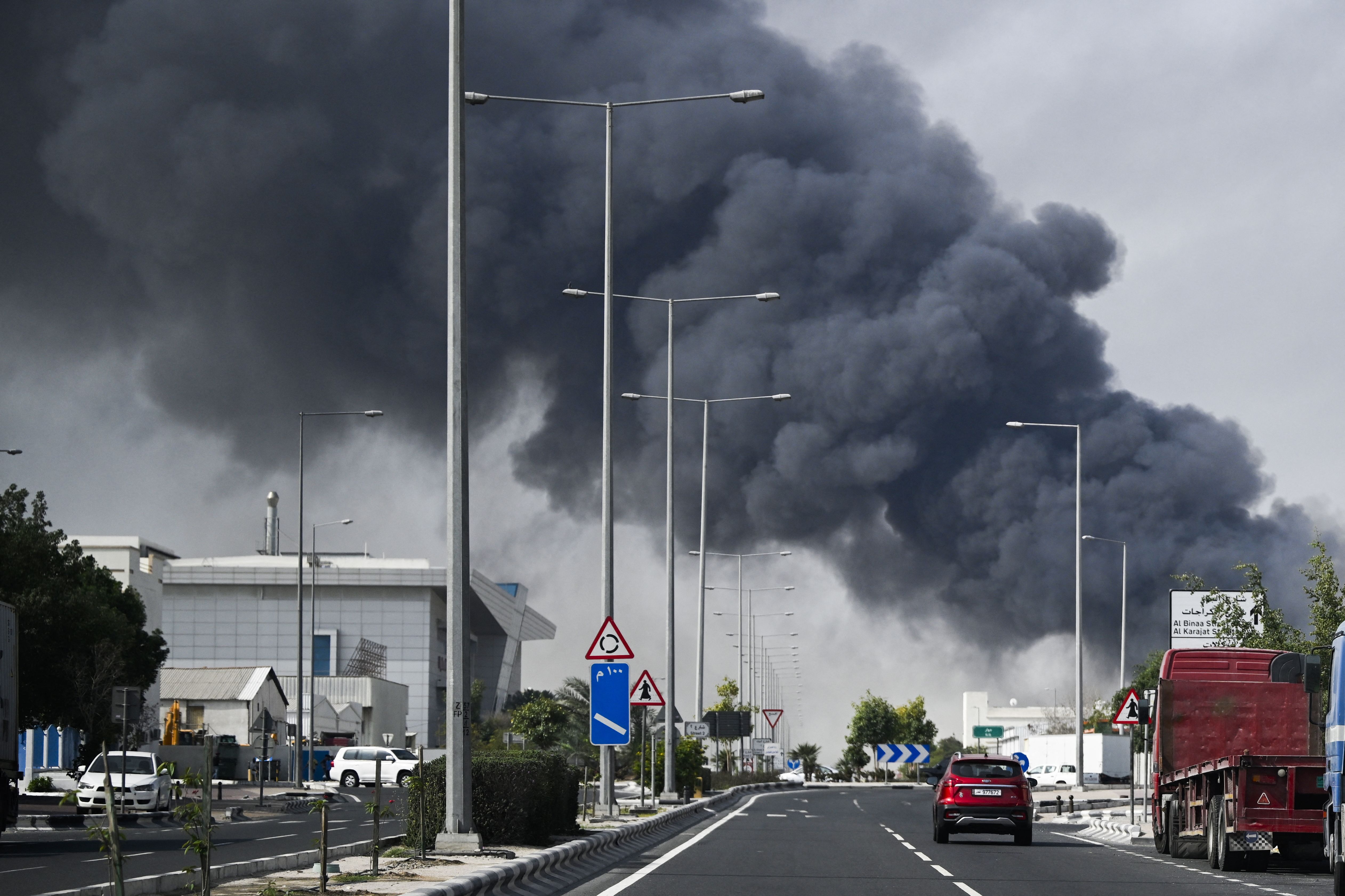 Cars driving on a highway with thick black smoke rising over industrial buildings in Doha after reported Iranian strikes.
