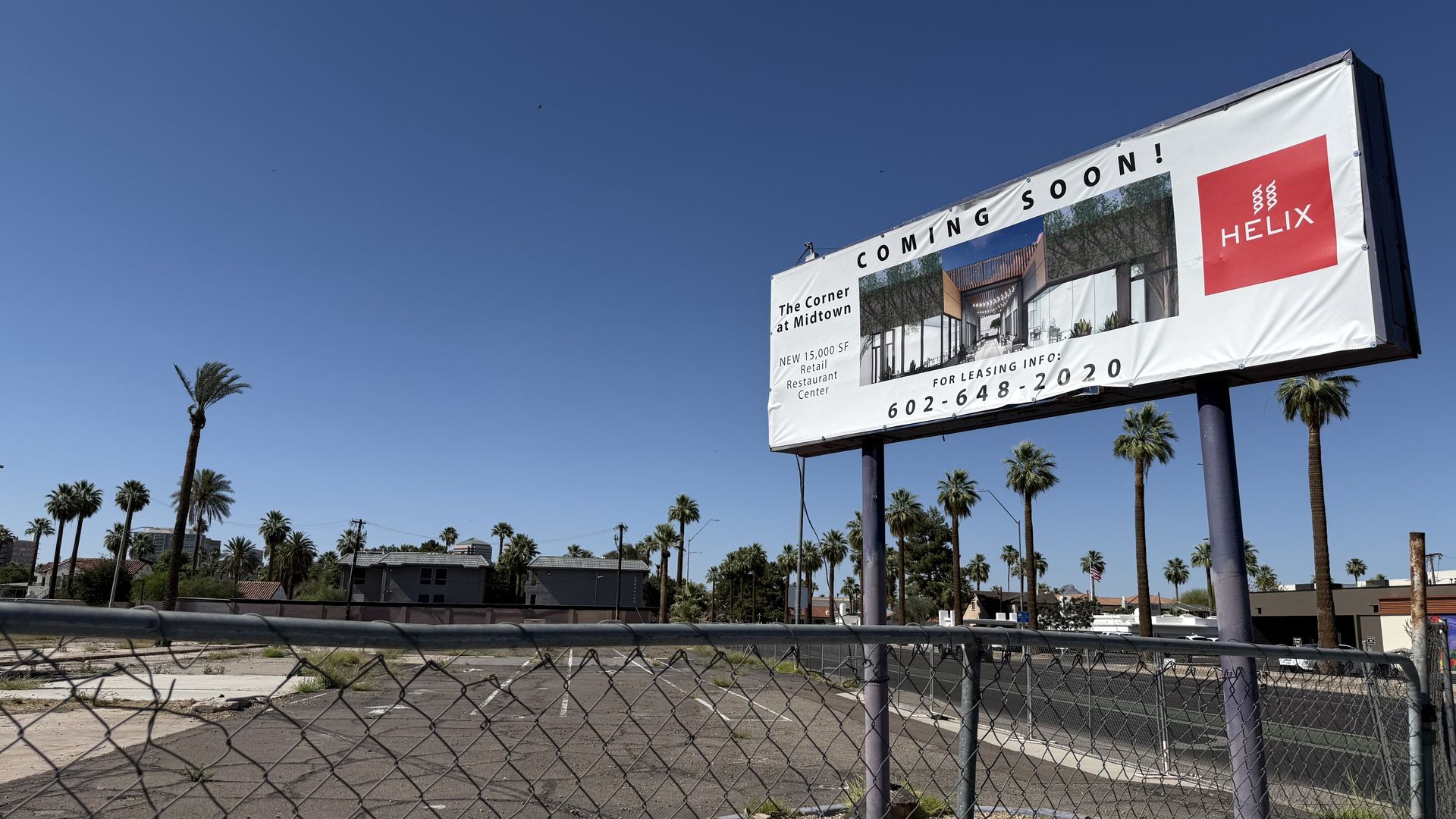 Large billboard on two purple poles against a blue sky, reading COMING SOON! with a red HELIX logo and a rendering of a retail center; the sign is in the corner of a vacant blacktop lot with palm trees in the middle, surrounded by a chain-link fence. 