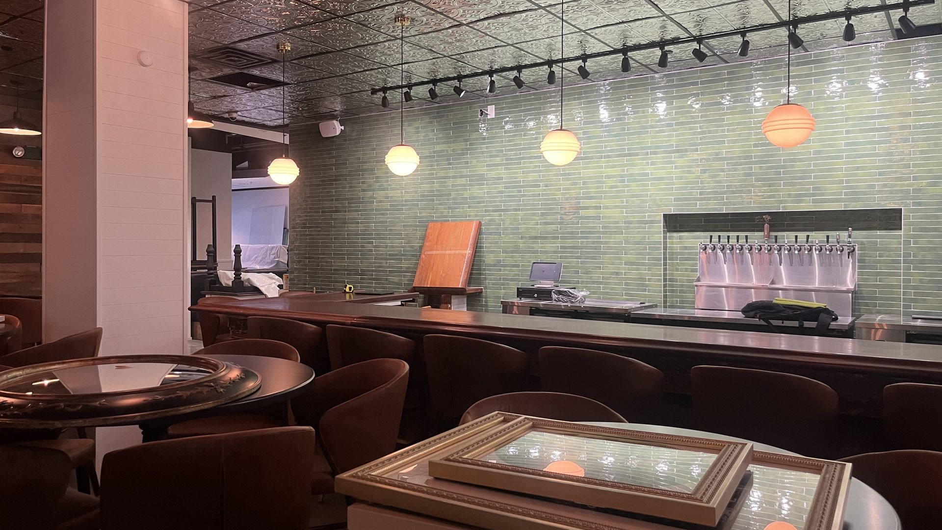 Interior of a cozy bar with brown chairs around tables, a long bar counter, green tile backsplash, round pendant lights, a white column on the left, and beer taps along the back wall.