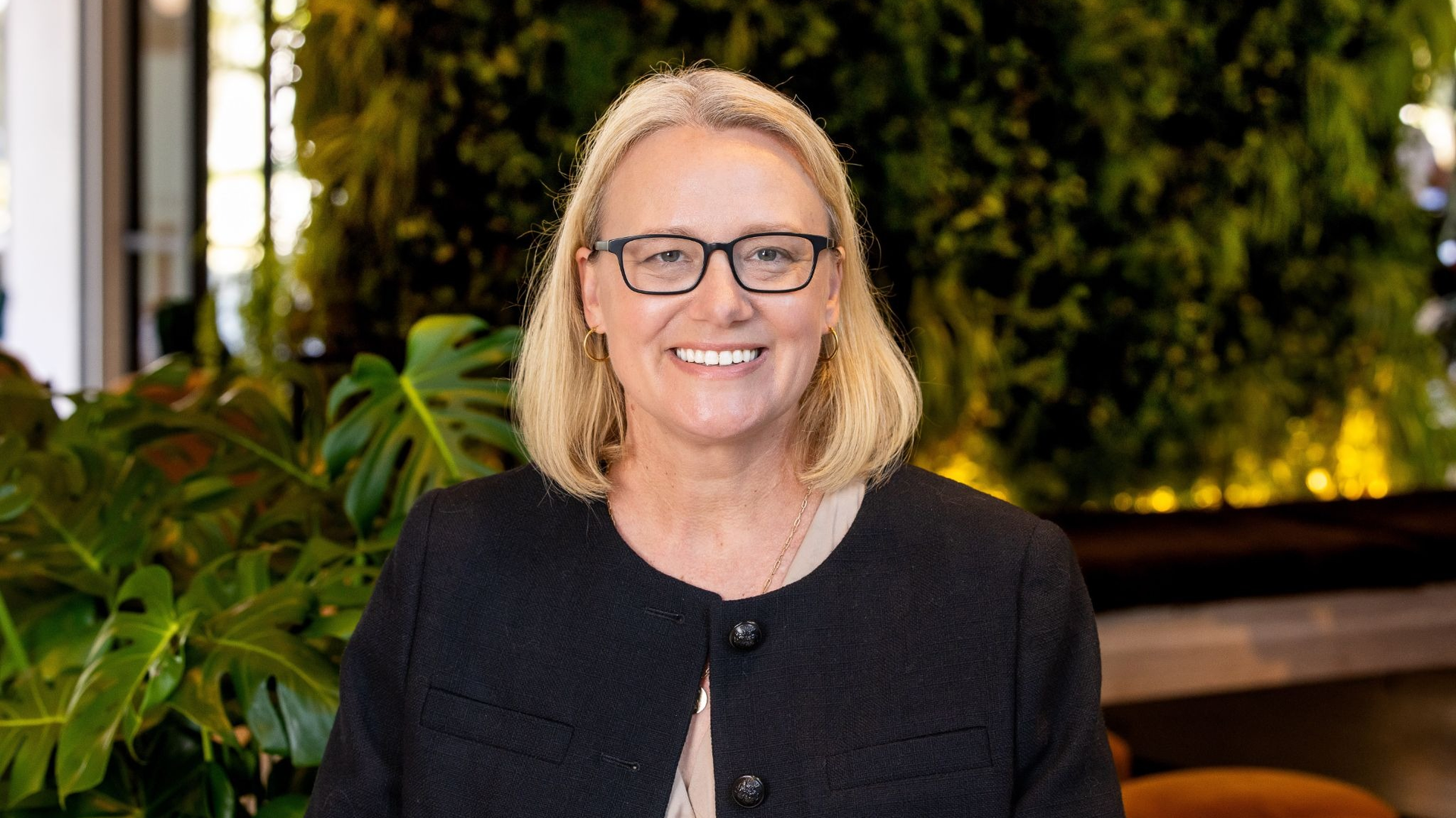 Smiling blonde woman with black-framed glasses and a black blazer posed in front of lush green plants.