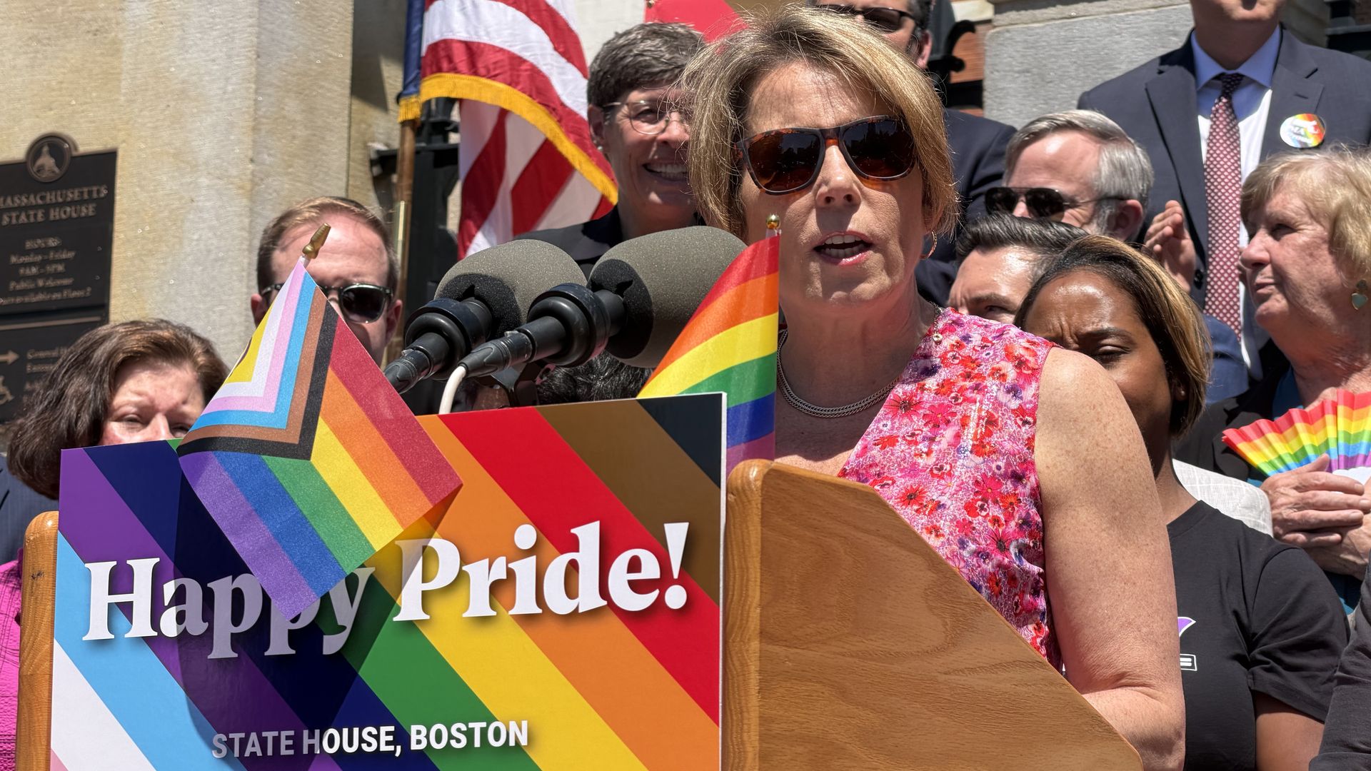 At a Pride event outside the Massachusetts State House in Boston, Gov. Maura Healey speaks from a lectern amid rainbow flags and a "Happy Pride!" sign on June 3, 2026. She's got sunglasses on because of the glaring sun. 