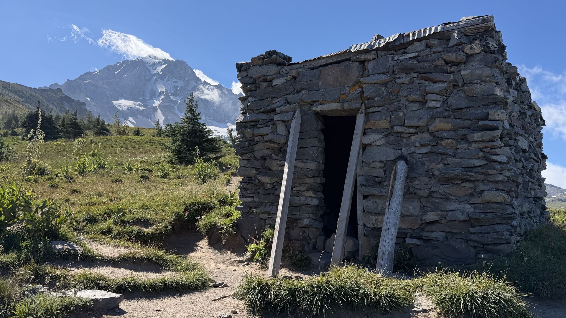 Stone hut with wooden supports on the right, open dark doorway; green alpine meadow with shrubs in foreground; snow-capped mountain peak and clear blue sky in background.