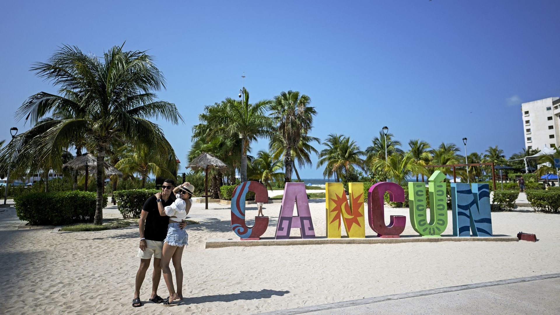 Sunny beach scene with palm trees, a colorful "CANCUN" sign, a couple posing in summer outfits, thatched umbrellas, and a bright blue sky over sandy ground and the ocean.