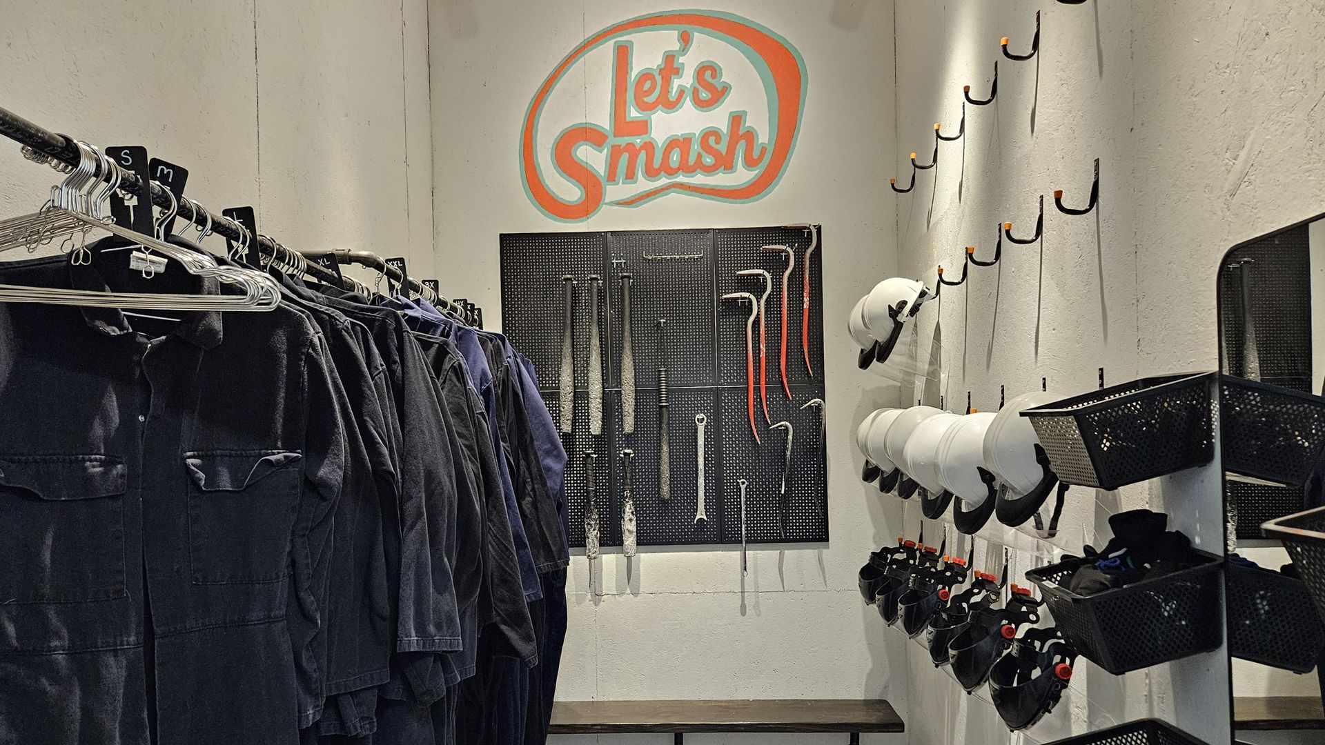 A back wall lined with weapons says "let's smash," with rows of coveralls and helmets on either side