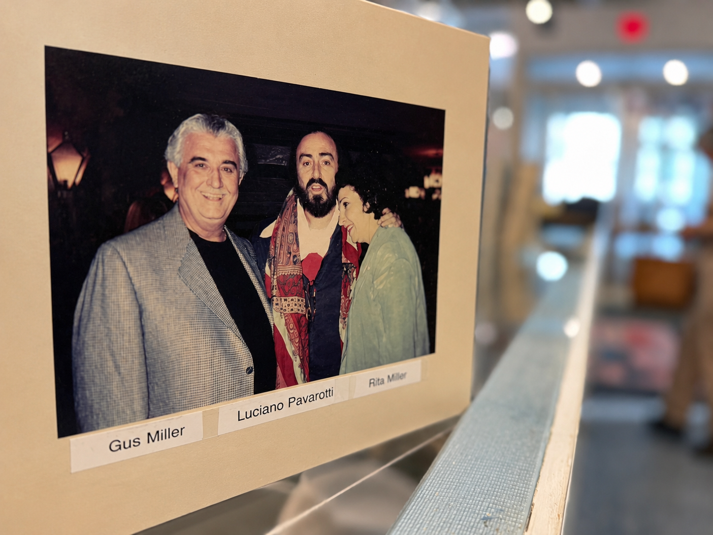 Framed photo shows three people posing: left older man in gray blazer, middle bearded man with scarf, right woman in teal jacket; labels read Gus Miller, Luciano Pavarotti, Rita Miller.
