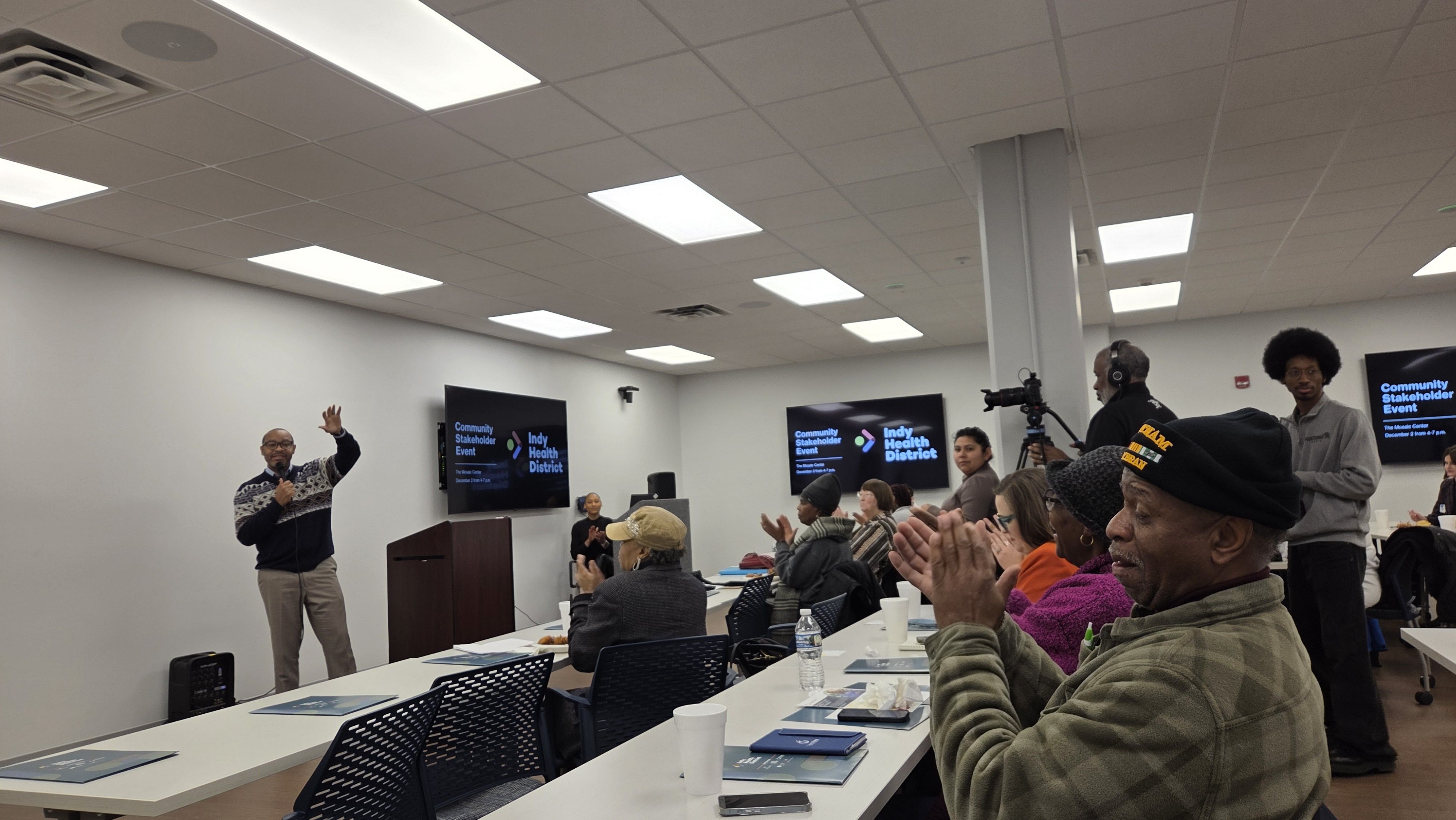 Man speaking with microphone in a room full of seated people applauding, with screens displaying "Community Stakeholder Event" and "Indy Health District" on the walls.