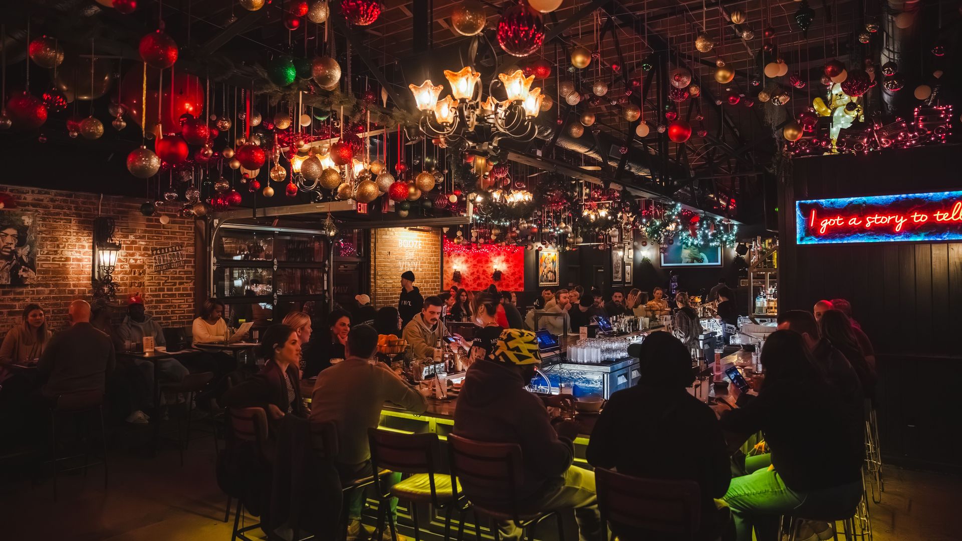 Cozy bar interior with red, gold, and white hanging ornaments, warm chandeliers, and neon sign reading I got a story to tell, patrons seated at bar and tables enjoying evening.