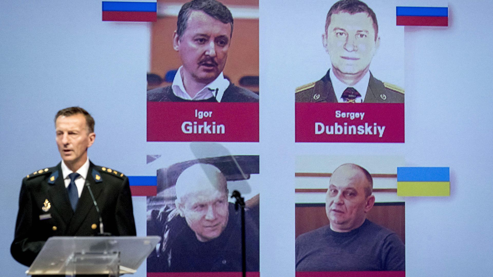 This image shows a man standing at a podium with posters of four men behind him.