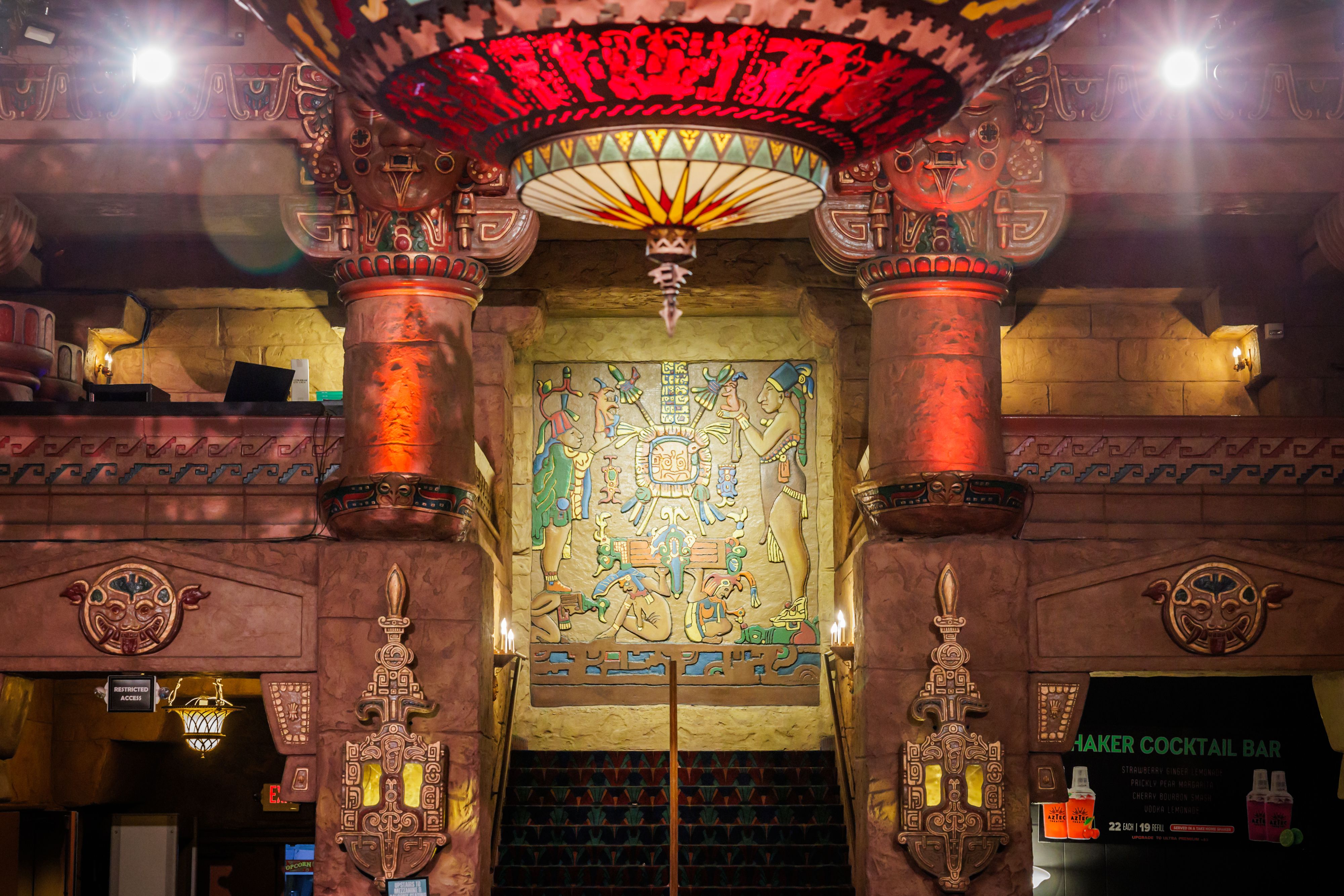 Interior scene with tall, carved stone pillars, a large colorful circular lantern hanging from the ceiling, and a mural of stylized figures on stone walls, bathed in warm lighting.