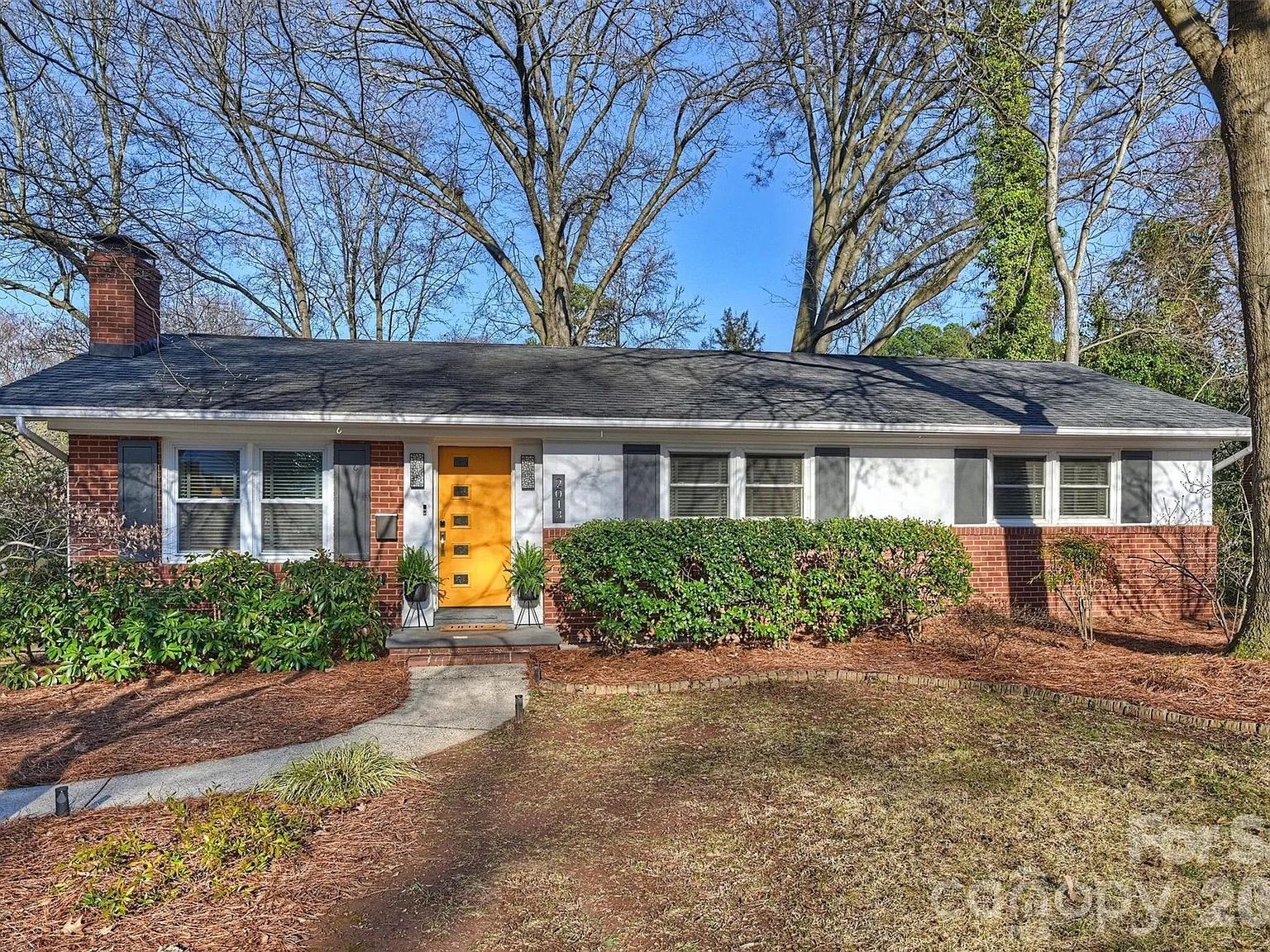 Single-story brick house with a bright yellow front door, grey shutters, a brick chimney, and bare trees in the background on a clear sunny day.