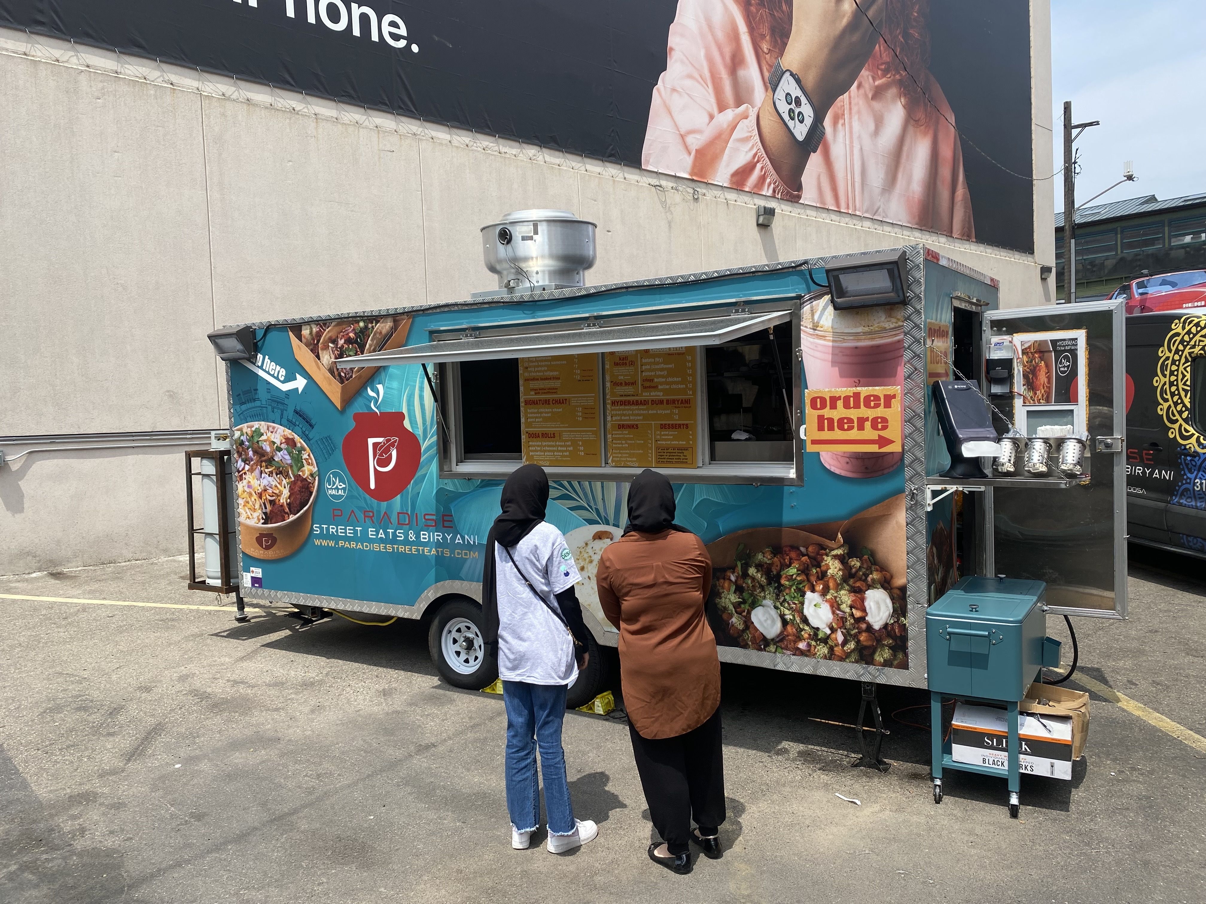 A blue food truck parked in a parking lot with two people in front of it deciding what to order.