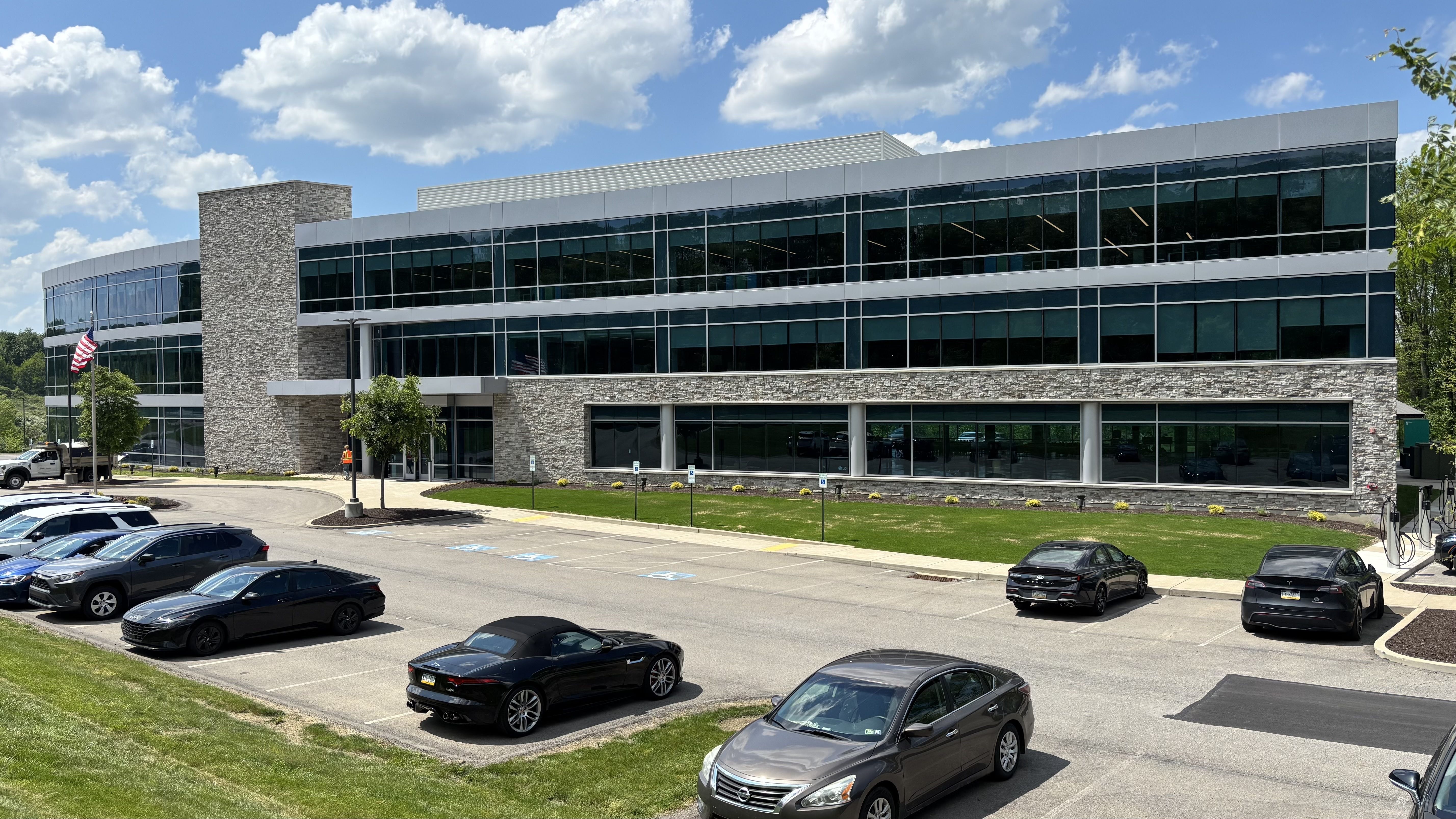 Modern two-story office building with glass façades, a stone base and entrance pillar, flagpoles with American flags, surrounding landscaping, and a parking lot full of cars under a blue sky.