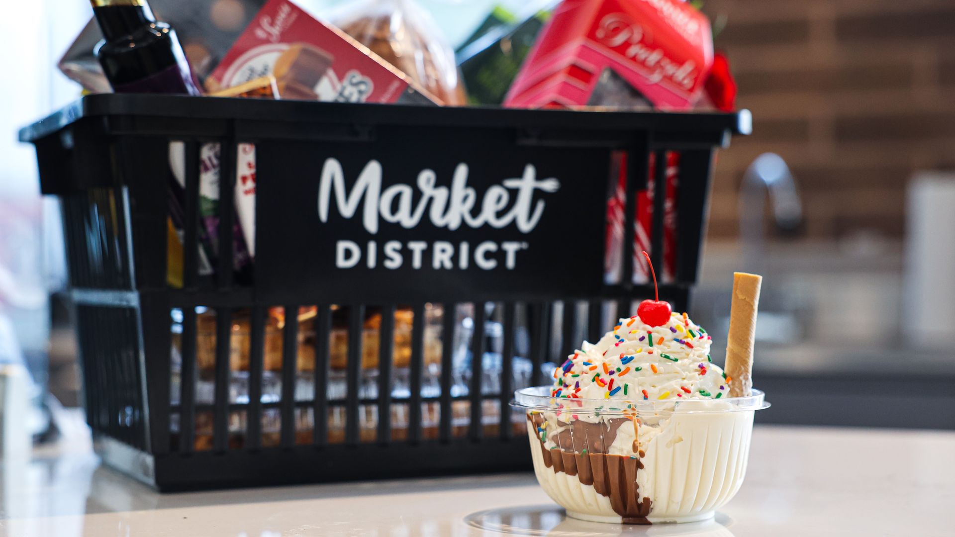 Ice cream in a bowl with whipped cream, rainbow sprinkles and a cherry sits on a white counter in front of a black Market District crate filled with snacks; a blurred kitchen backdrop.