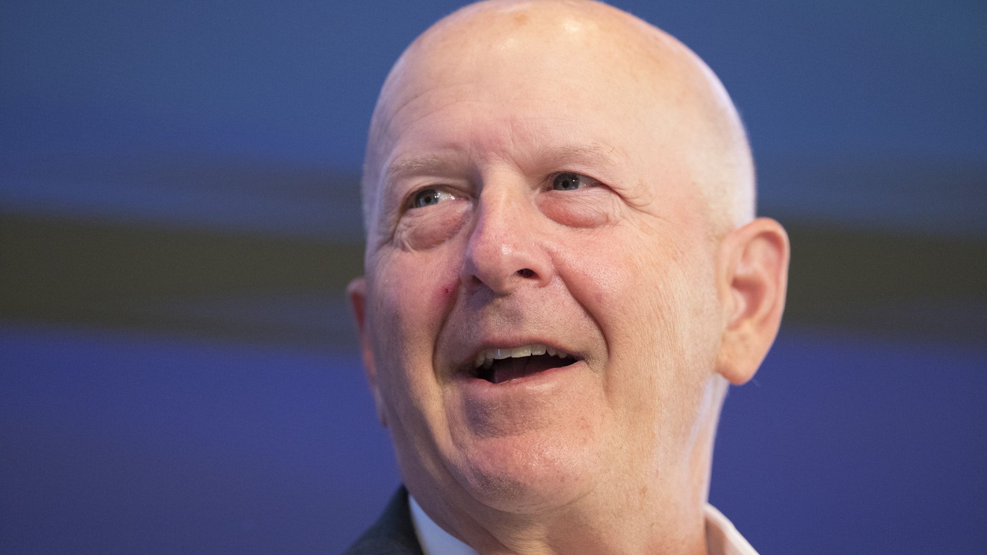 A person with a shaved head speaks in front of a deep blue background