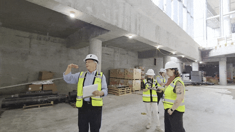 A group of construction workers wearing white helmets and yellow safety vests inside a large unfinished concrete building, discussing plans with papers and devices.