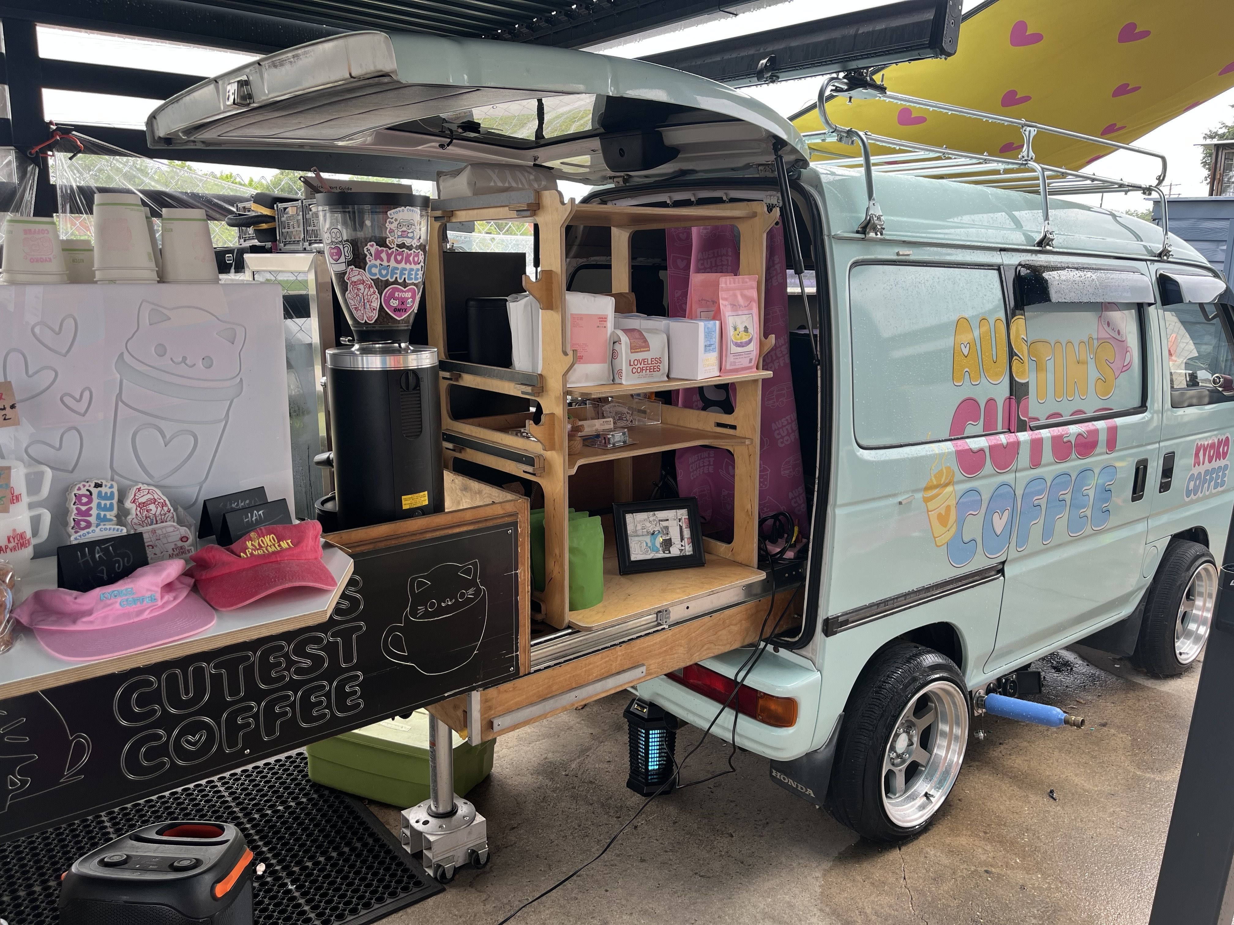 Light blue vintage van converted into a mobile coffee shop with an open side displaying wooden shelves, cups, a blender, and pink tote bags; yellow canopy with pink hearts above.