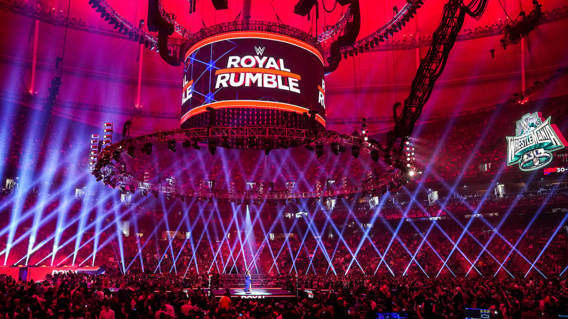The inside of a stadium packed with people with a Jumbotron screen reading "ROYAL RUMBLE."  