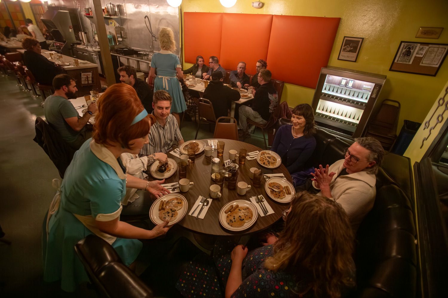 Busy retro diner with people seated around a round table, a waitress in a turquoise uniform serves dishes; warm yellow walls with orange panels, a kitchen in view and a beverage cooler in back.
