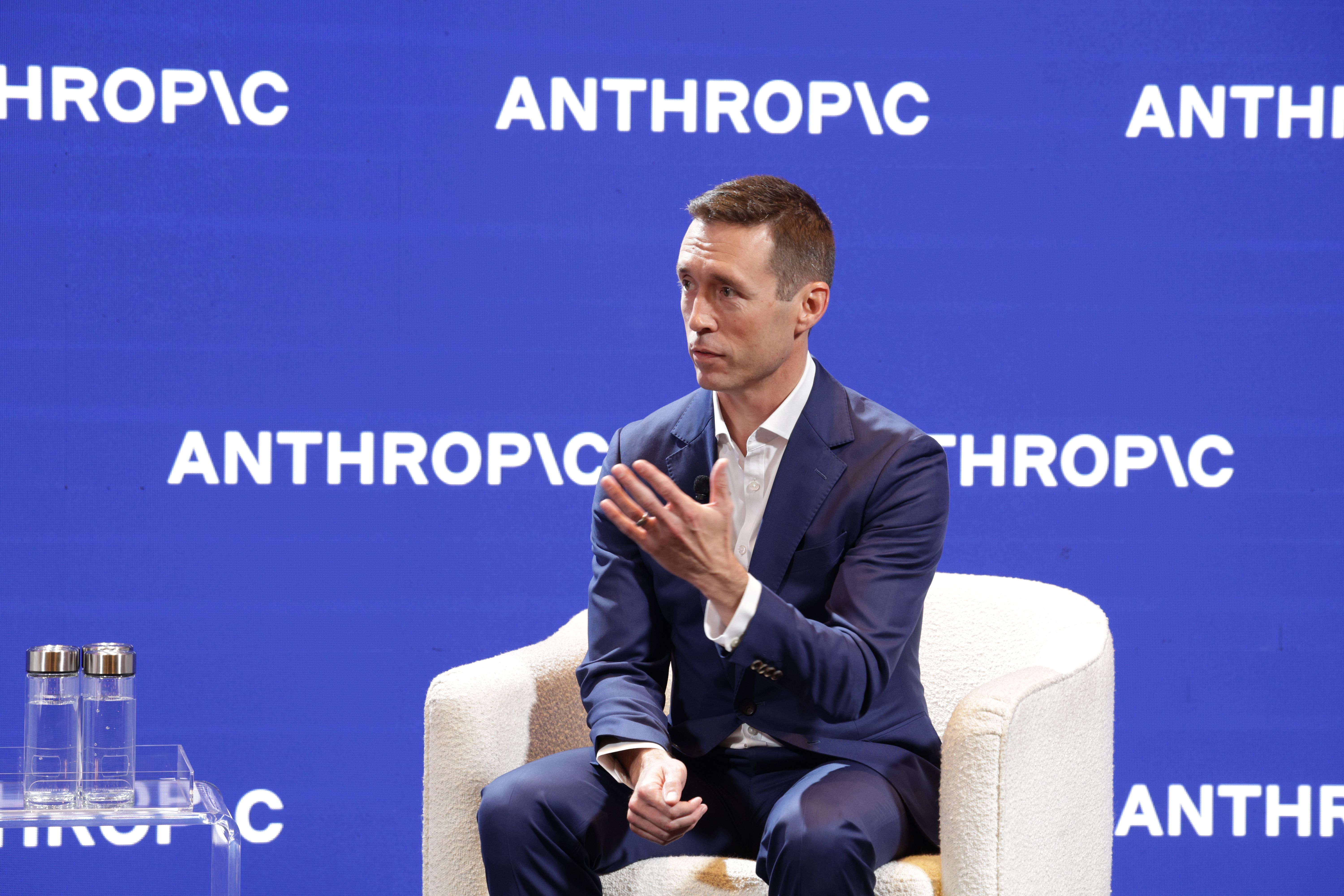 A man in a navy suit sits on a white chair during a panel against a blue backdrop repeating the word "ANTHROPIC". He gestures with his hands; glass water bottles sit on a clear table nearby.