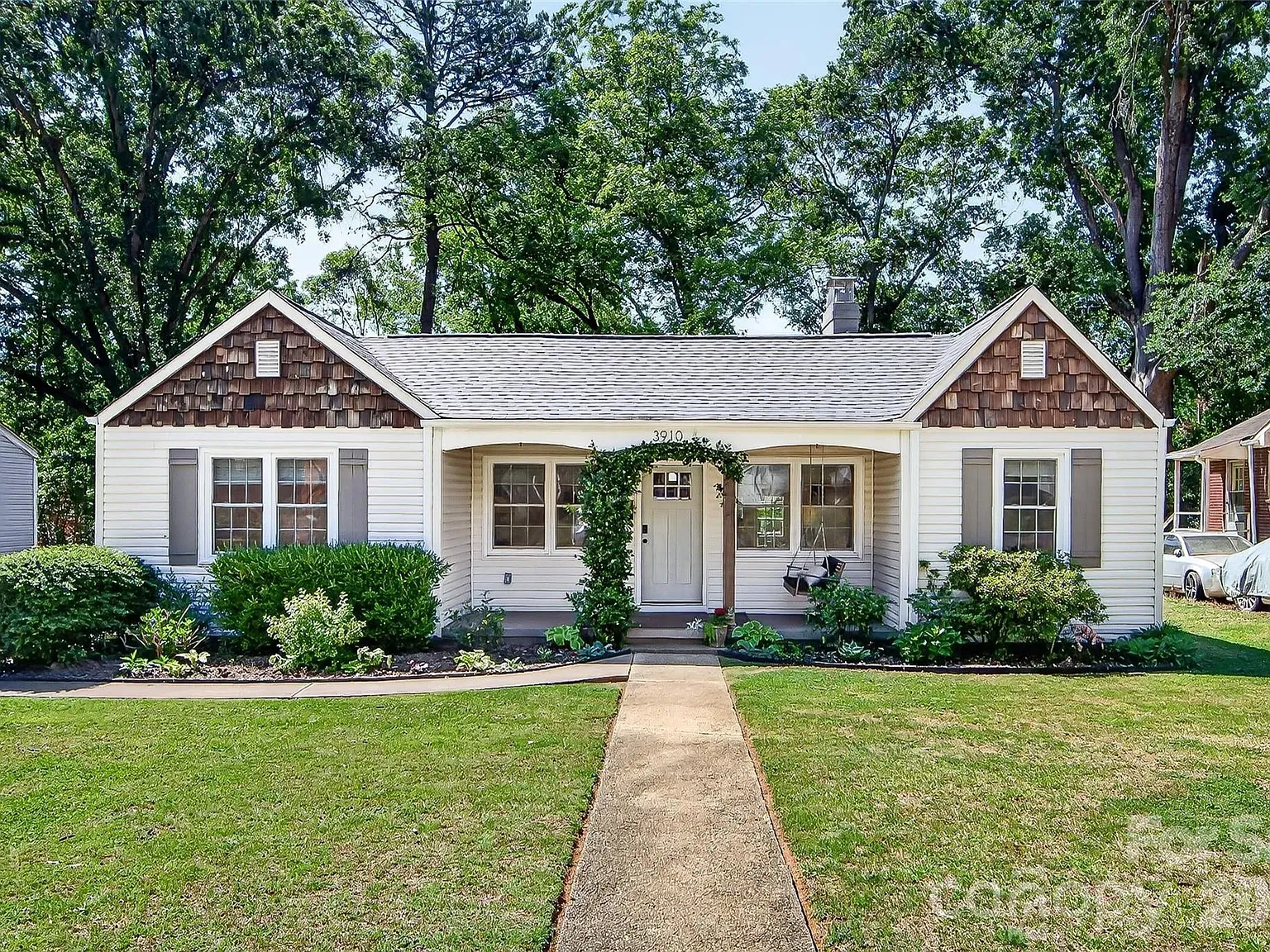 Single-story white house with brown shingle gables, a central front door under a small covered porch, and a leafy ivy arch. Well-kept lawn, shrubs, and tall trees behind.