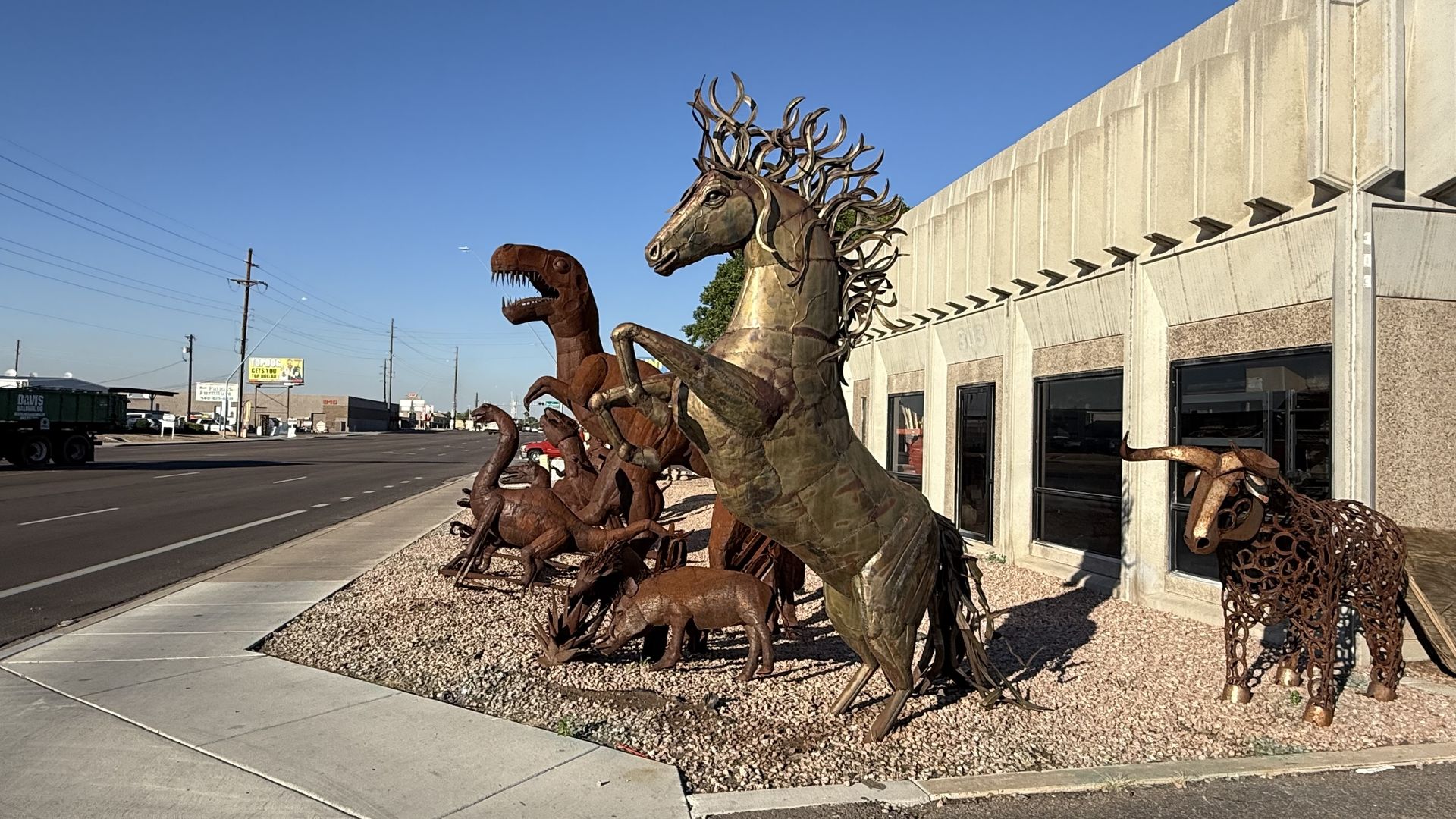 Rusted metal sculptures of dinosaurs and a rearing horse line a gravel bed beside a beige concrete building on a sunny street under a blue sky.
