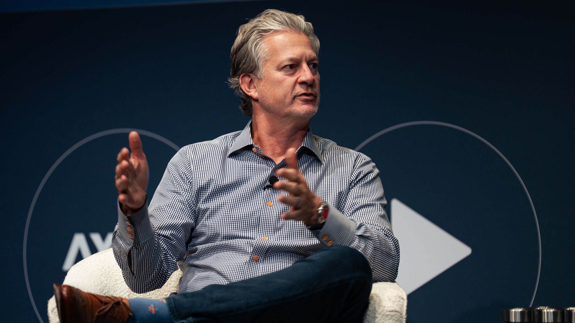Neil Vogel sits in a beige chair on a stage, wearing a checkered shirt and dark jeans. He gestures with his hands while speaking against a dark blue backdrop.