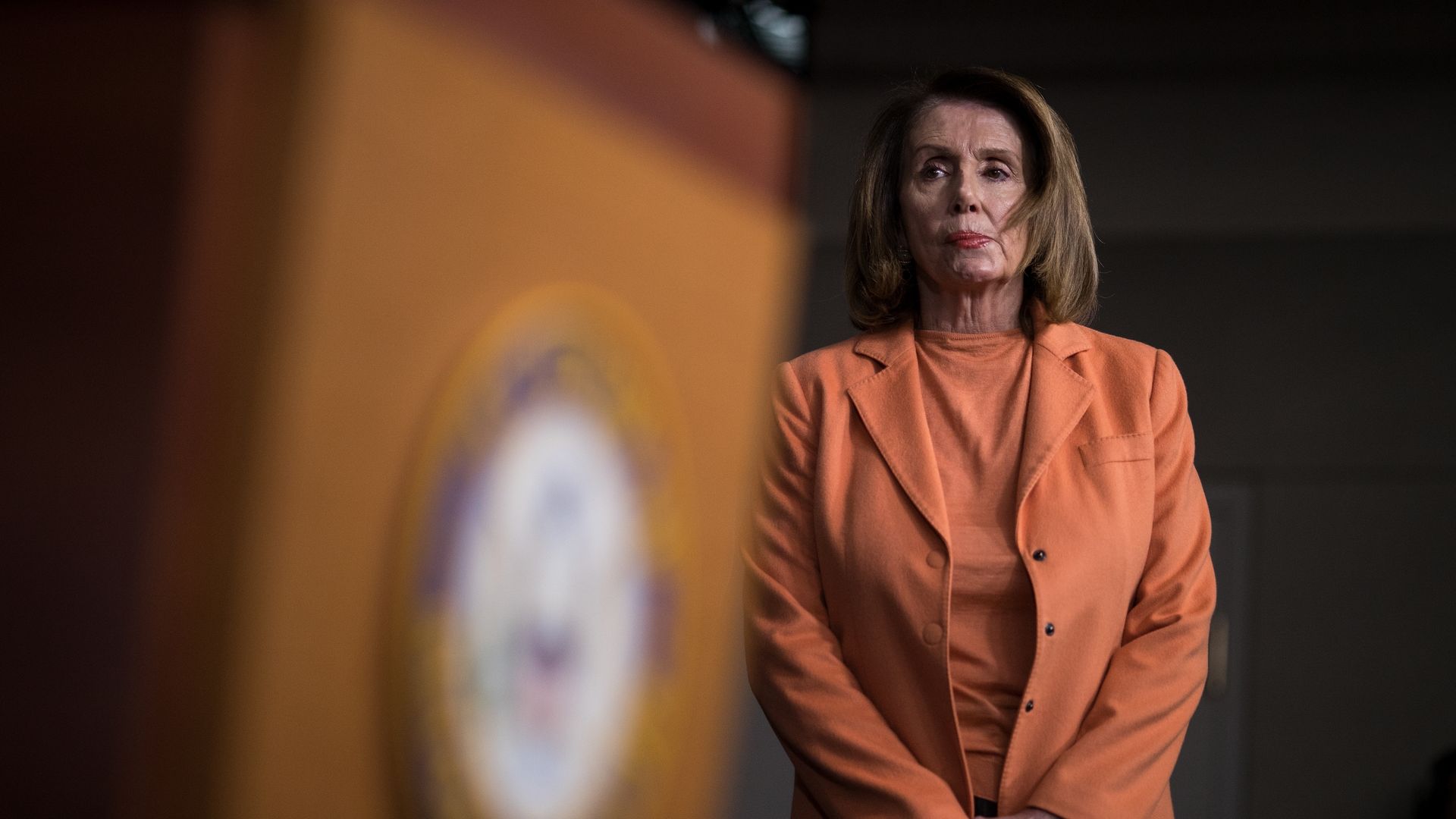 Nancy Pelosi standing next to a podium in an orange suit.