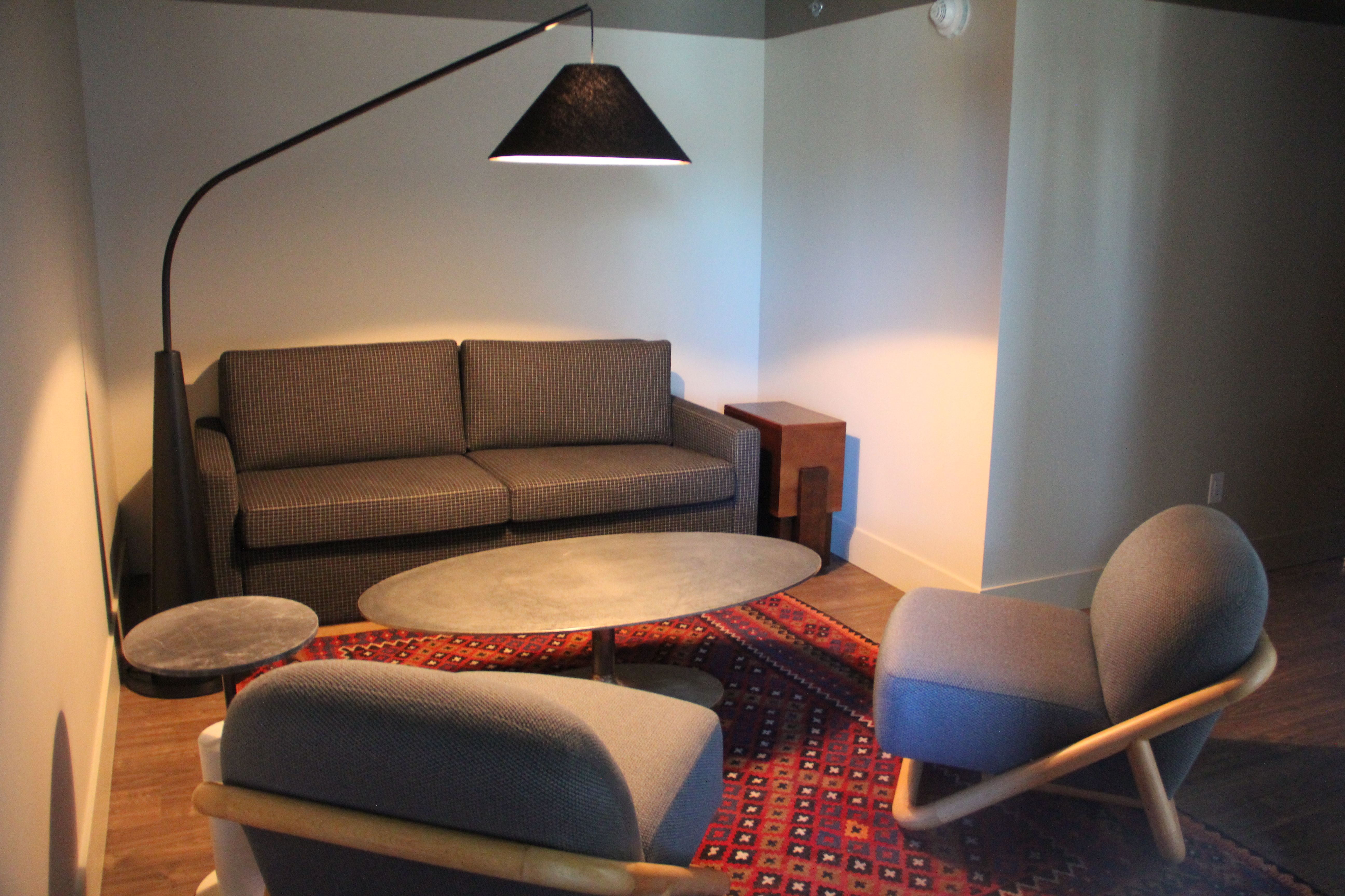 A cozy living room with a gray check sofa, two blue-gray armchairs, a round marble coffee table, a curved black floor lamp, a small side table, and a red patterned rug.