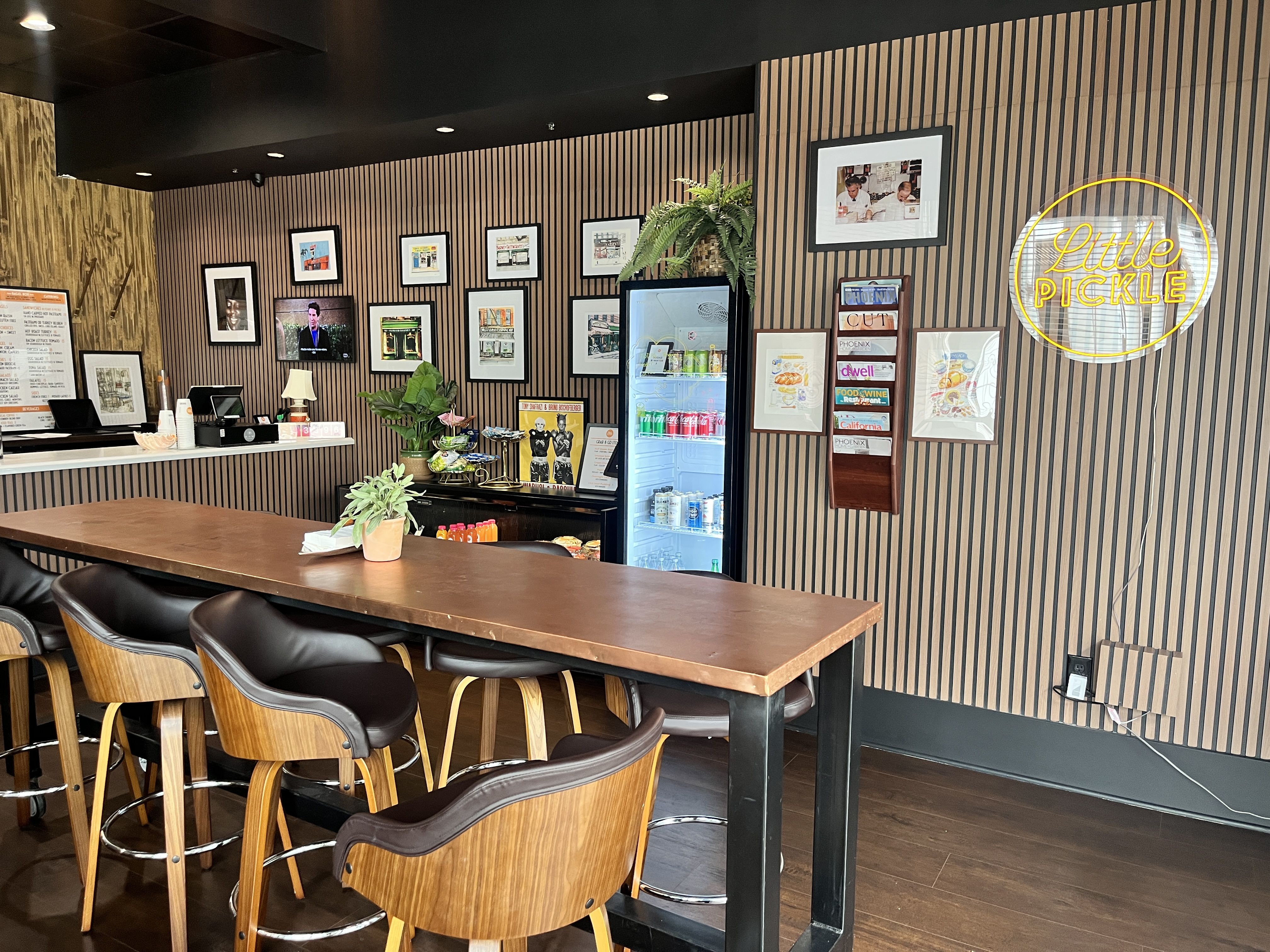 Interior of Little Pickle restaurant with wooden high chairs, a long wooden table, striped walls, framed pictures, a fridge with drinks, and a yellow neon sign reading "Little Pickle".