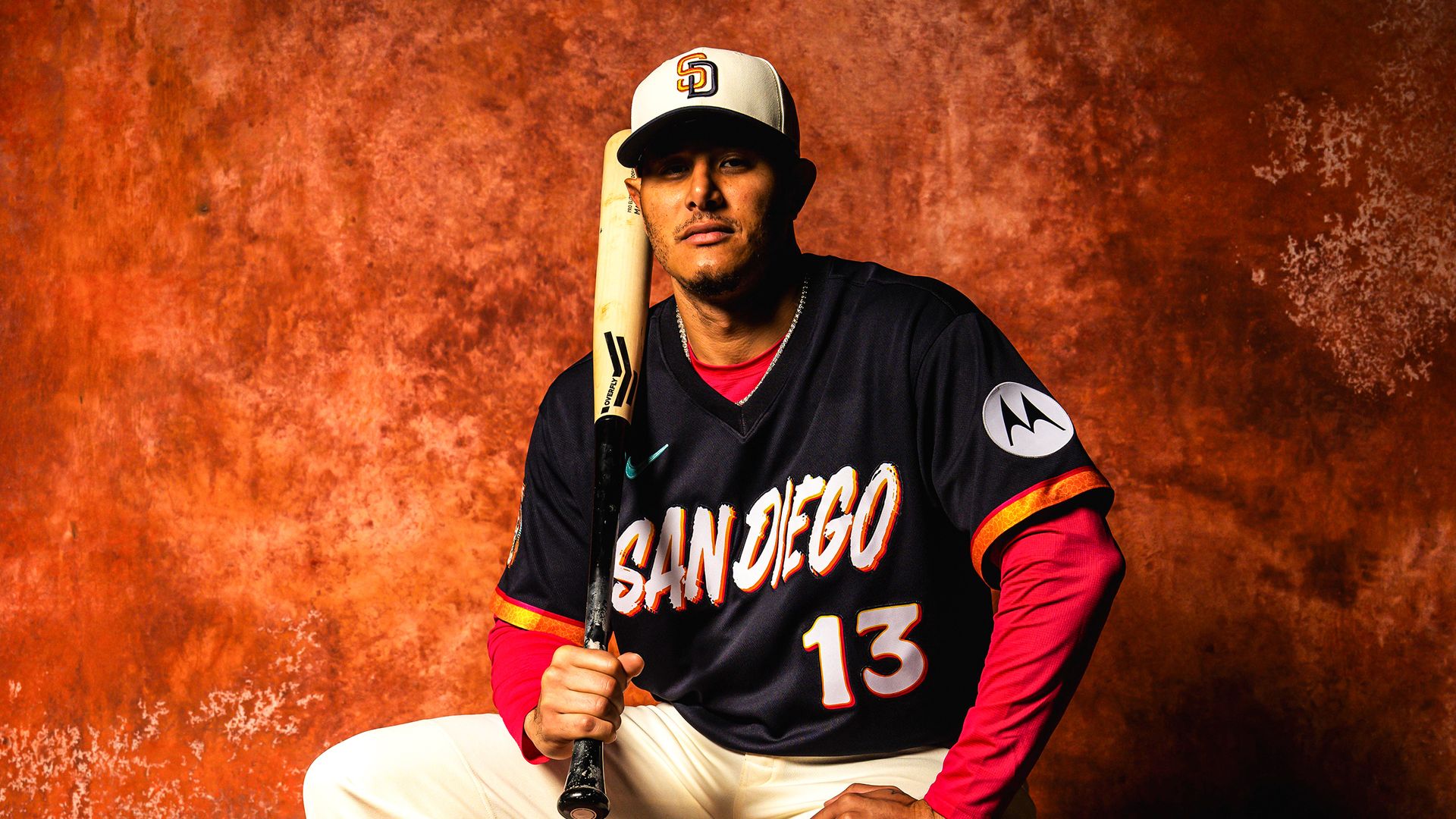 Manny Machado wears a dark navy San Diego Padres jersey with the number 13 and red sleeves sits against a textured rust-orange background, holding a wooden bat over his shoulder.