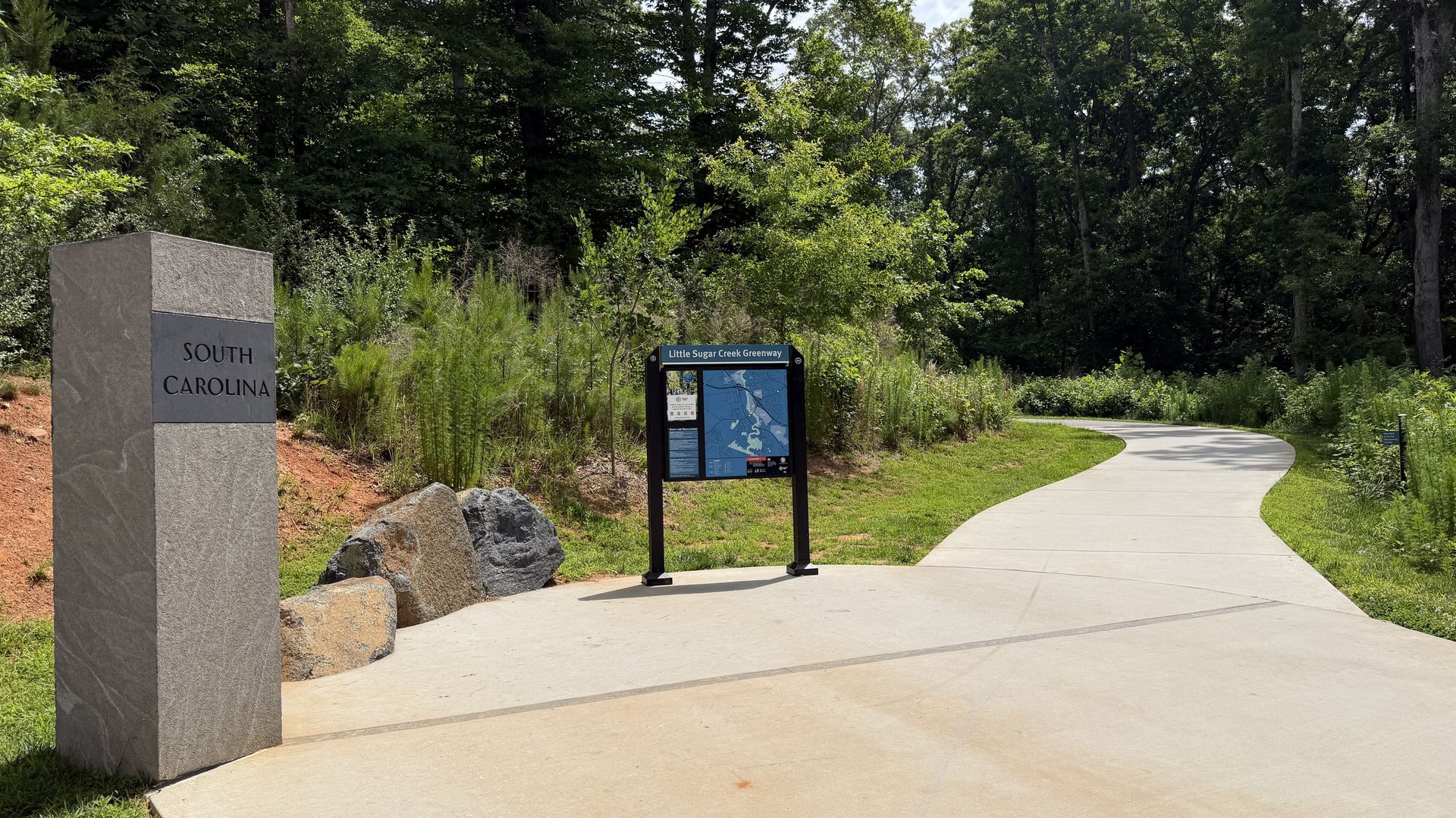 Sunlit park entry with a tall gray stone pillar reading "South Carolina" on the left, a map sign labeled "Little Sugar Creek Greenway" and a curved concrete path winding through lush trees.