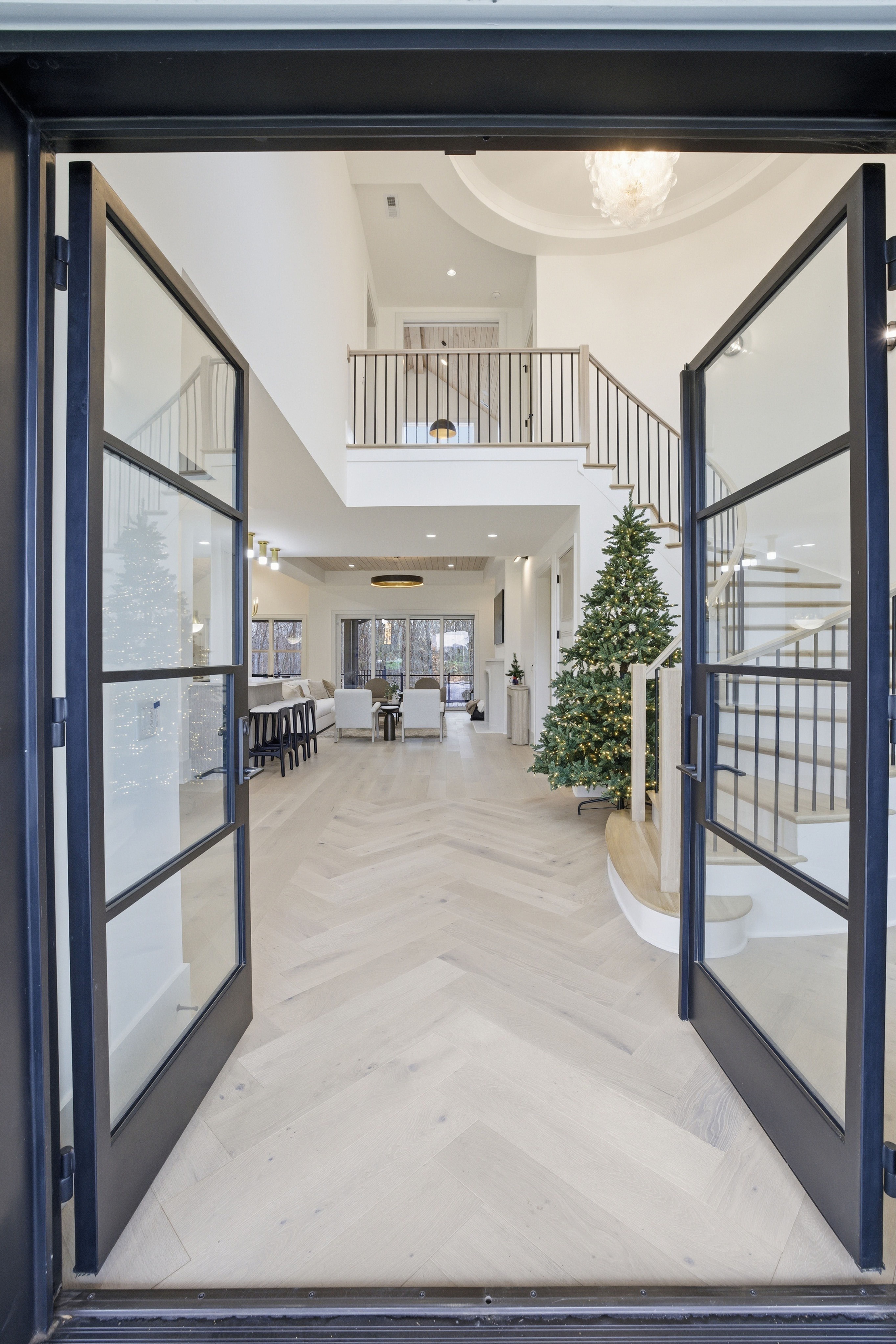 Open black-framed glass doors lead into a bright, modern living room with white walls, light chevron wood floors, a lit Christmas tree near stairs, and a second-floor balcony.
