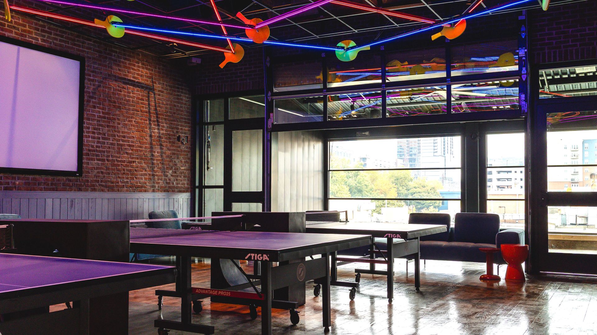 Indoor room with black Stiga ping pong tables, brick walls, large windows, colorful neon lights and ping pong paddles hanging from ceiling, and modern seating area.