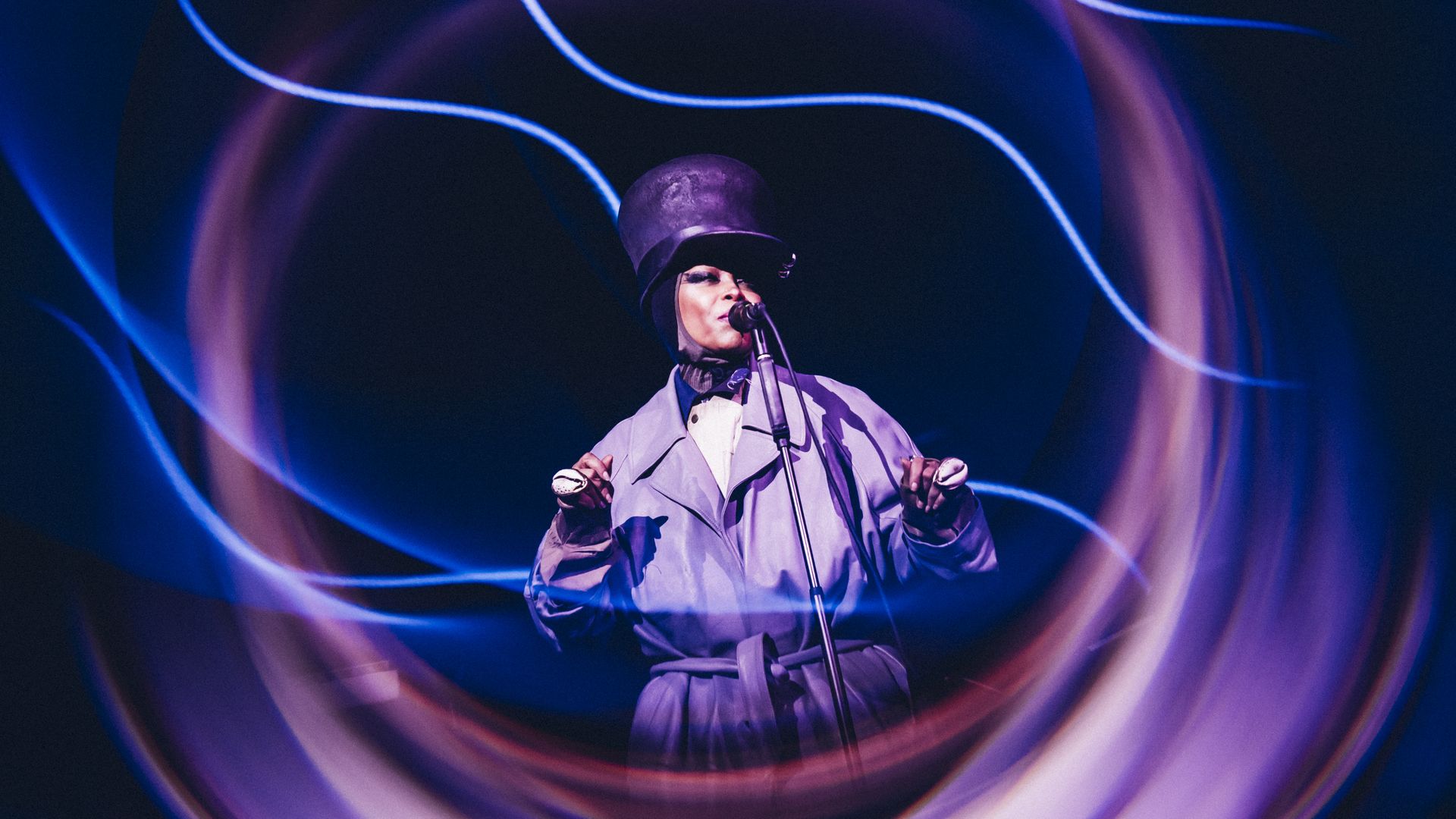 A performer wearing a tall black hat and a light gray coat sings into a microphone on stage, surrounded by swirling blue and purple light trails that form circular patterns.