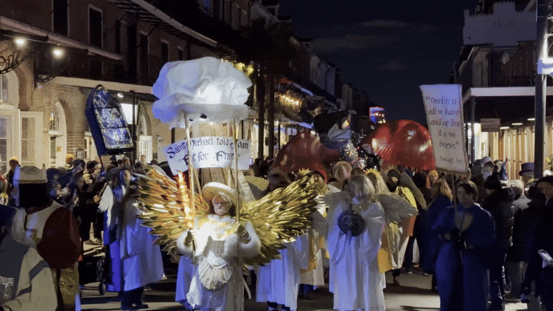 Night parade with people dressed as angels with glowing golden wings and holding signs, walking through a street lined with brick buildings and onlookers.