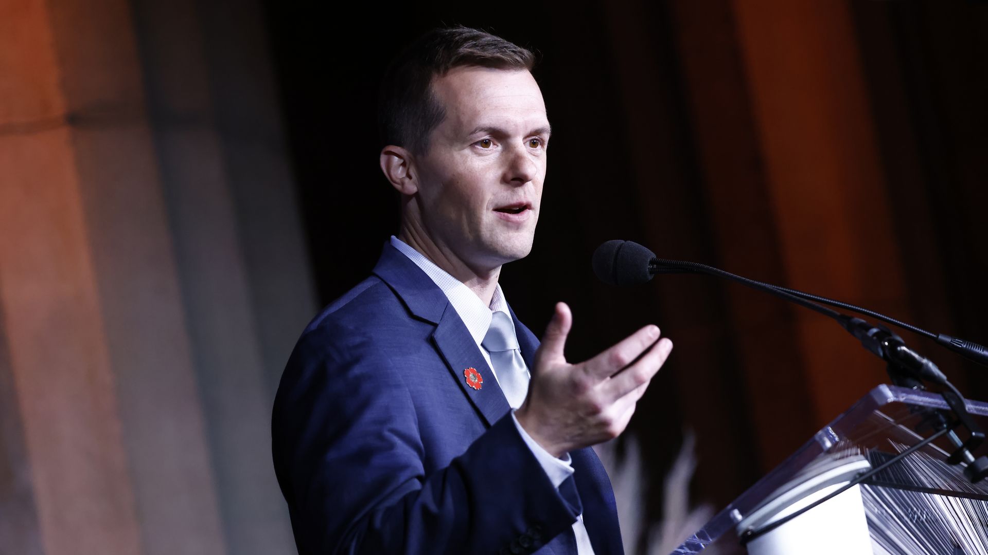 Rep. Jared Golden, wearing a blue suit and speaking into microphones at a glass podium in front of marble columns.