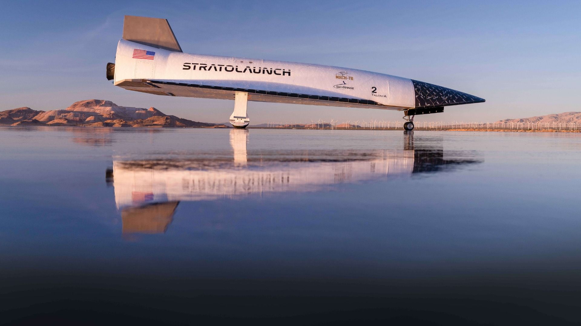 White Stratolaunch aircraft rests on a reflective salt flat, with desert mountains and distant wind turbines on the horizon under a blue sky.