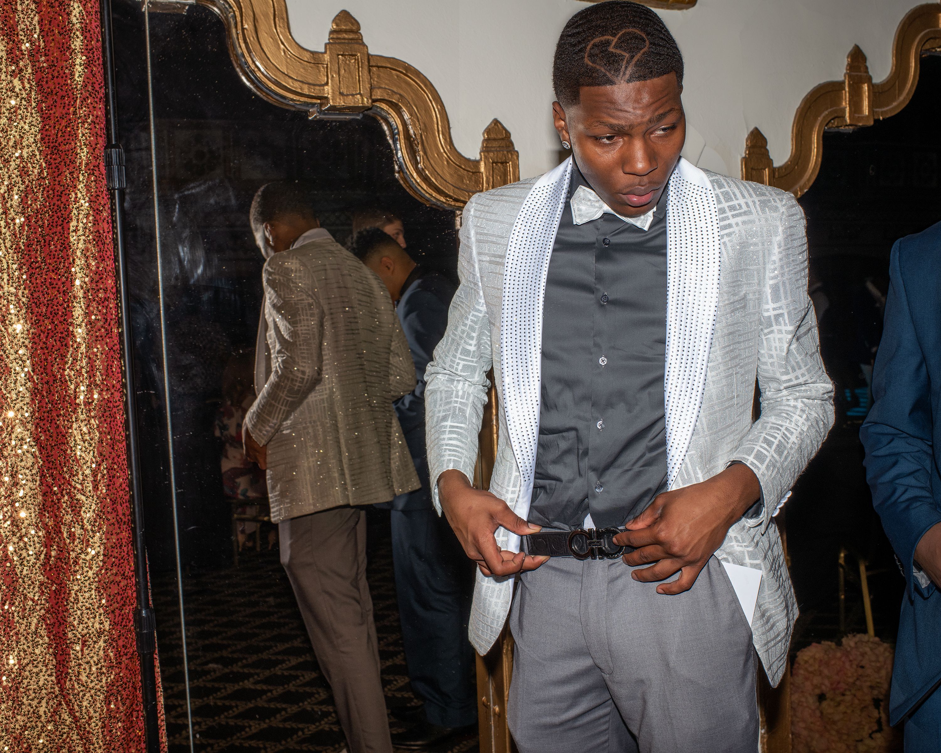 Young man in patterned white tuxedo jacket with embellished lapels, black shirt, bow tie, and gray pants adjusting belt. He has a heart haircut design on his head. Mirror and red gold curtain background.