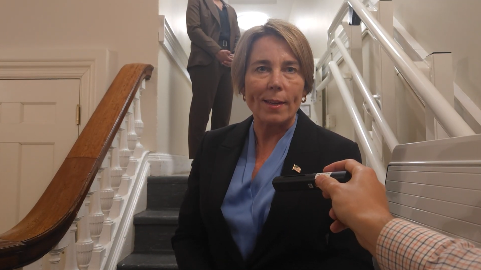 Woman in a black blazer and blue blouse sits on stairs, being interviewed. A hand holds a recorder toward her as a man stands at the top of the stairs.