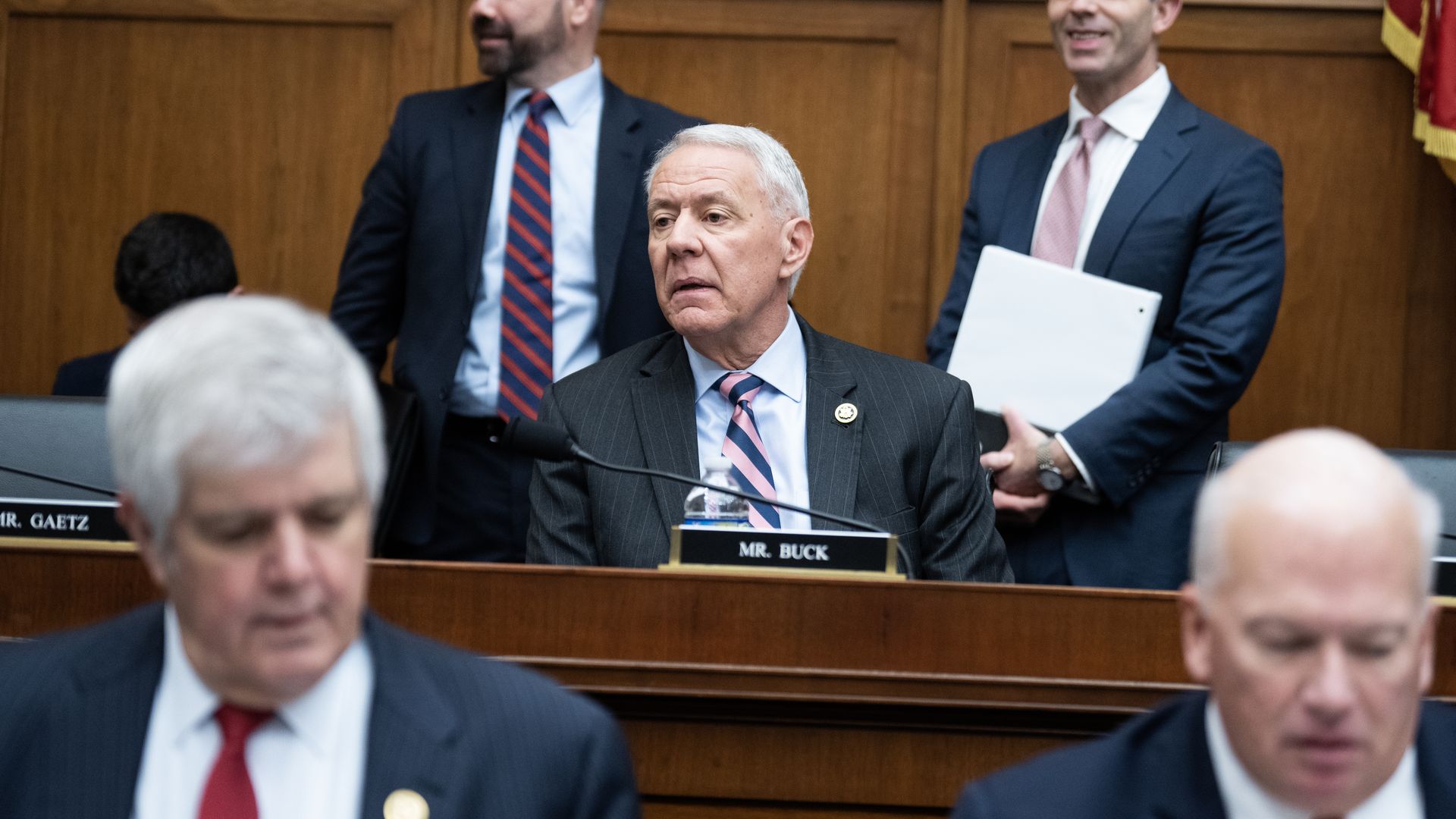 Rep. Ken Buck, wearing a dark gray suit, light blue shirt and blue and pink striped tie, sitting at a committee dais.