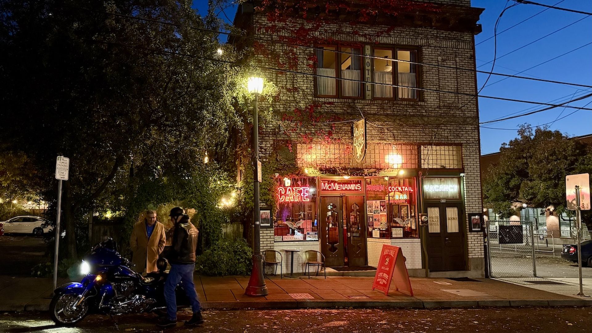 Evening scene of a brick building with neon signs reading "Cafe," "McMenamins," "Cocktail Bar," and "Vacancy" with two people near a blue motorcycle under a streetlamp.