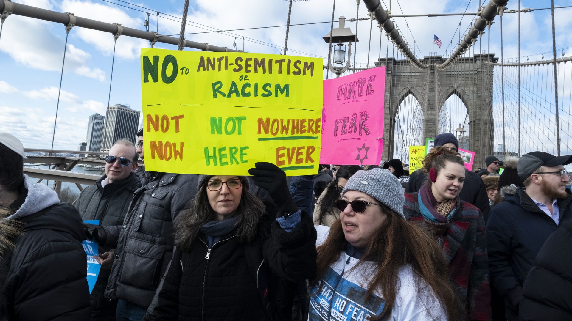 A marcher walking across the Brooklyn Bridge carrying a sign of "No to Anti-semitism or Racism Not Now, Not Here Nowhere Ever"