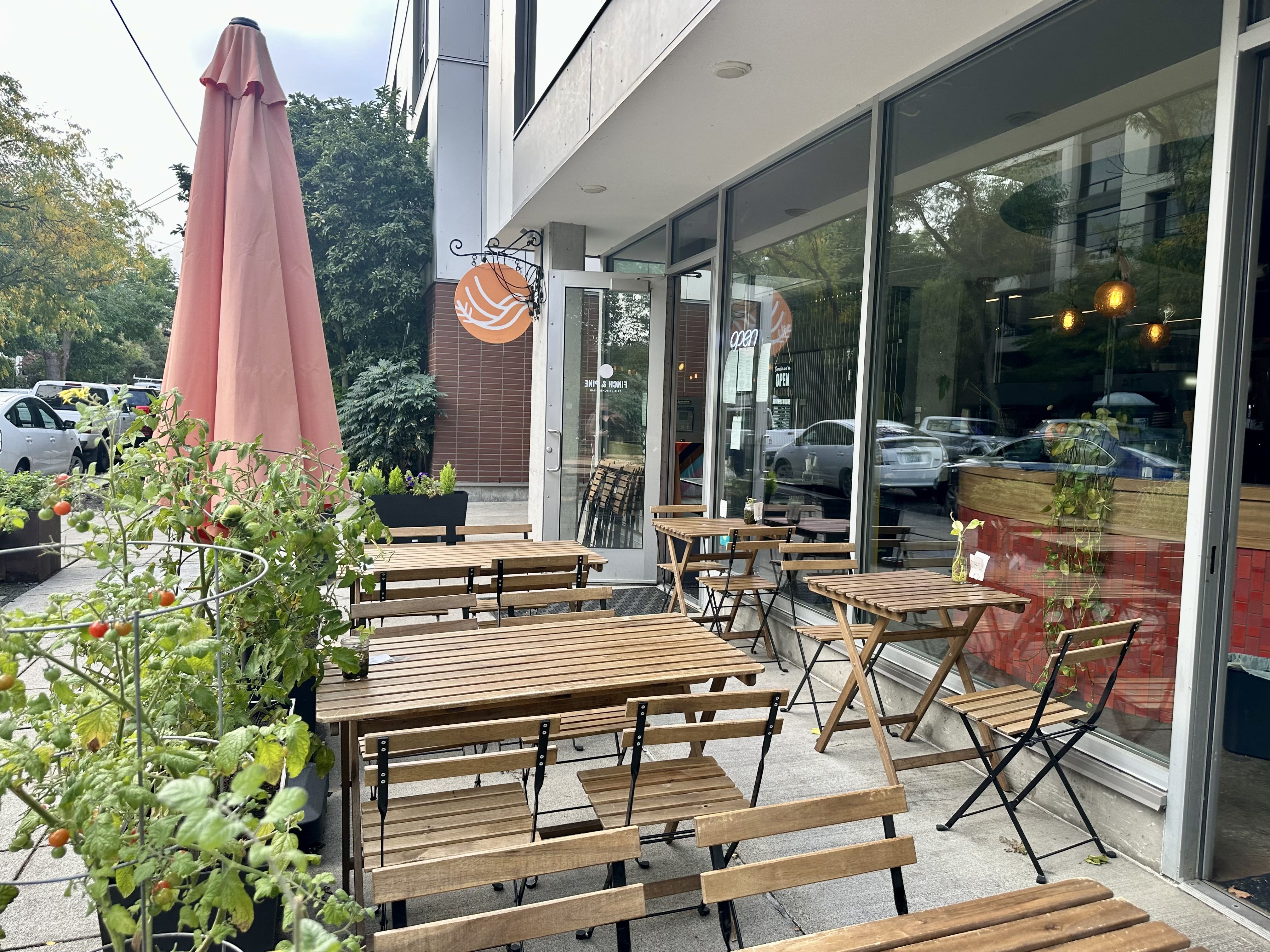 Outdoor tables and a closed umbrella in front of a restaurant with large windows and a circular sign that has a design featuring a bird on it.