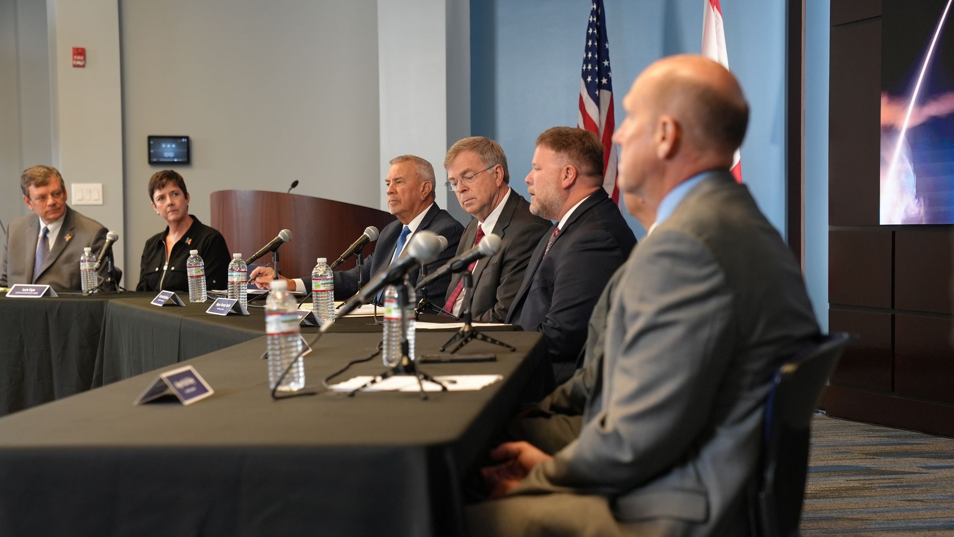 Six people in formal attire sit at a long black table with microphones and water bottles, against a blue wall with American and another flag, in a conference or panel setting.