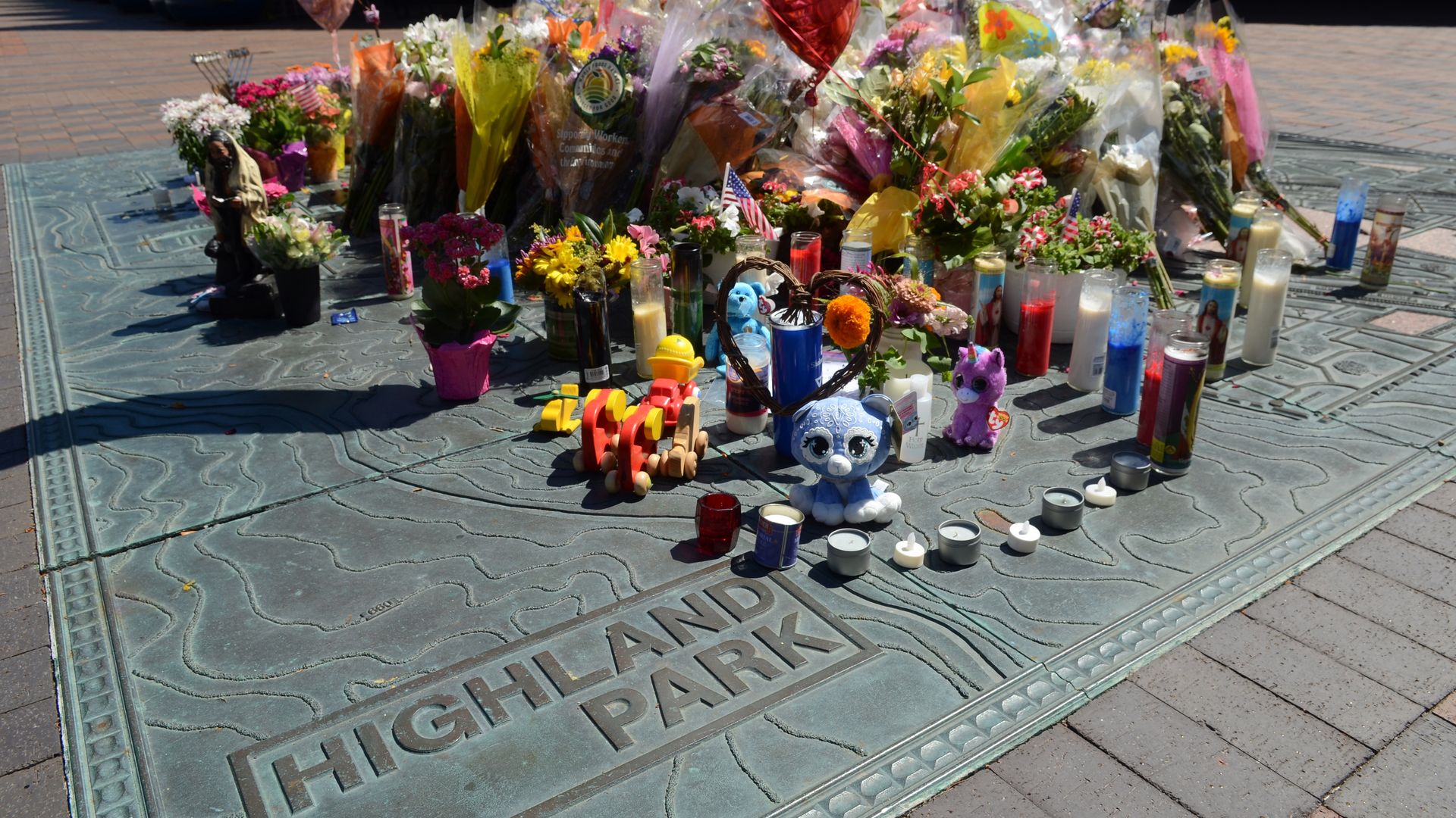 Photo of flowers and stuffed animals at a memorial site with a plaque that reads "Highland Park"