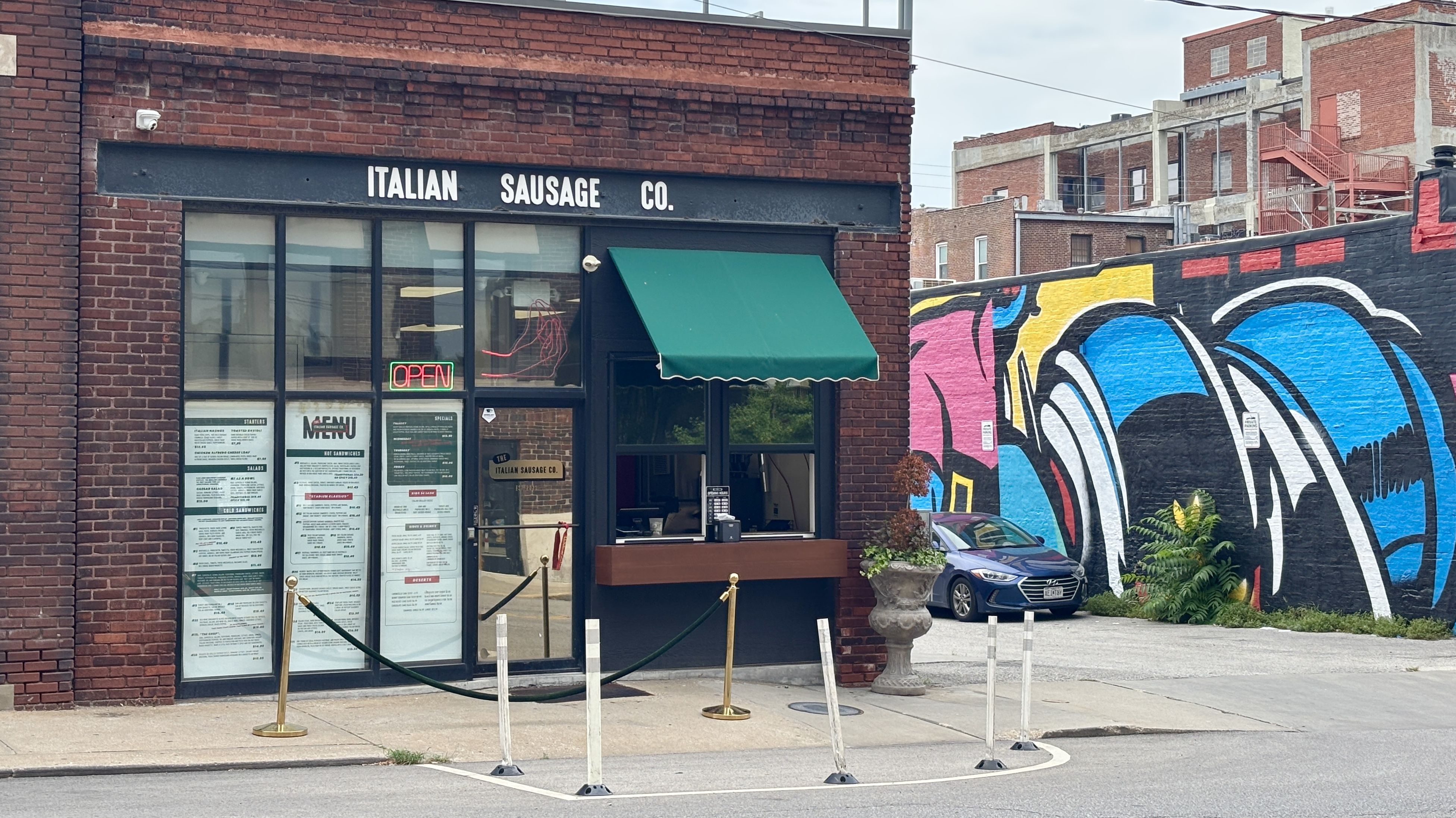 Brick storefront with a dark sign reading "ITALIAN SAUSAGE CO." and a green awning; large windows display menus and a neon "OPEN" sign. Colorful mural on the adjacent wall and a parked car nearby.
