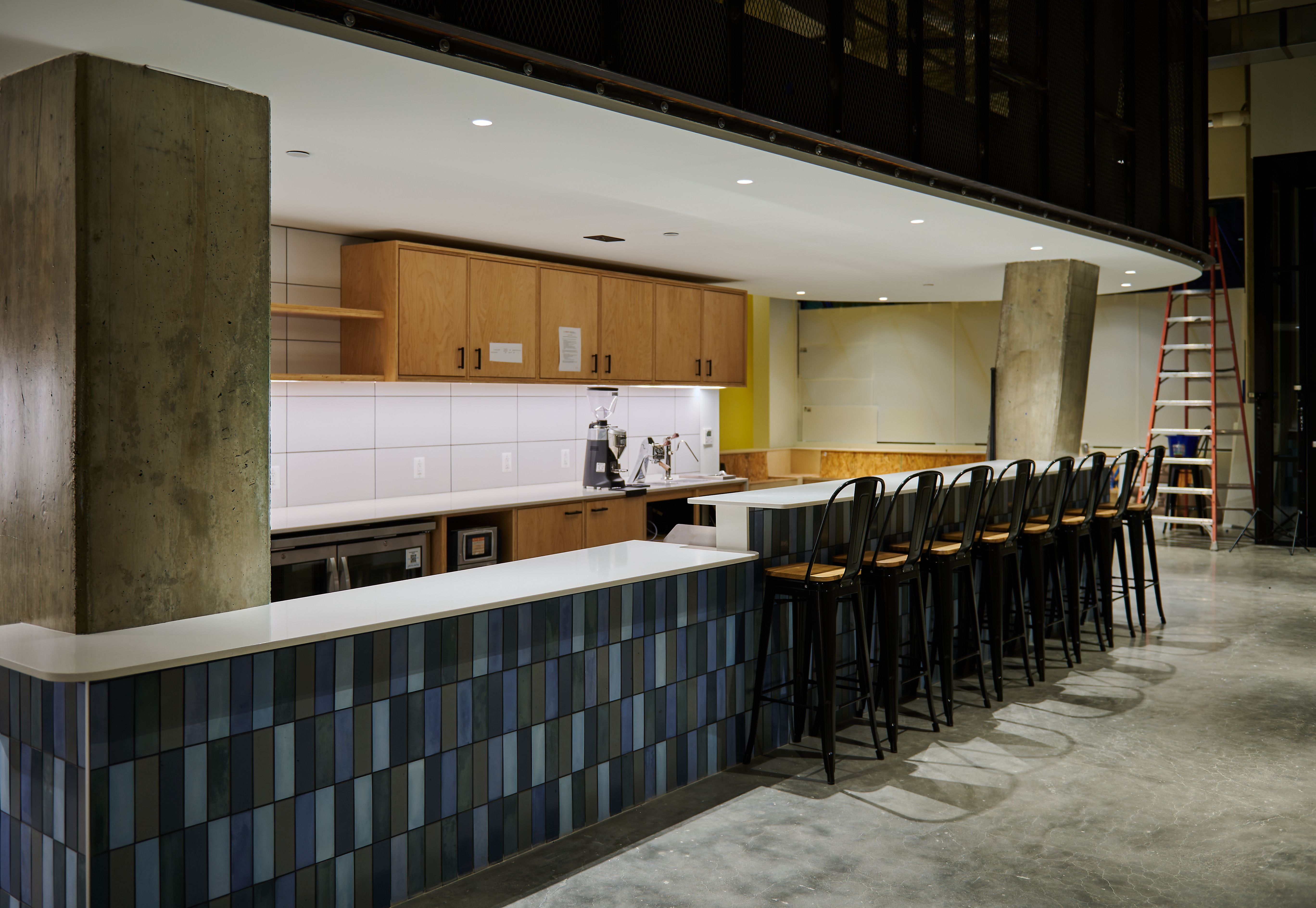 Modern cafe interior with a long white counter, blue tiled base, wooden cabinets, espresso machine, and black stools. Concrete columns and a mezzanine with railing above; a red ladder to the right.