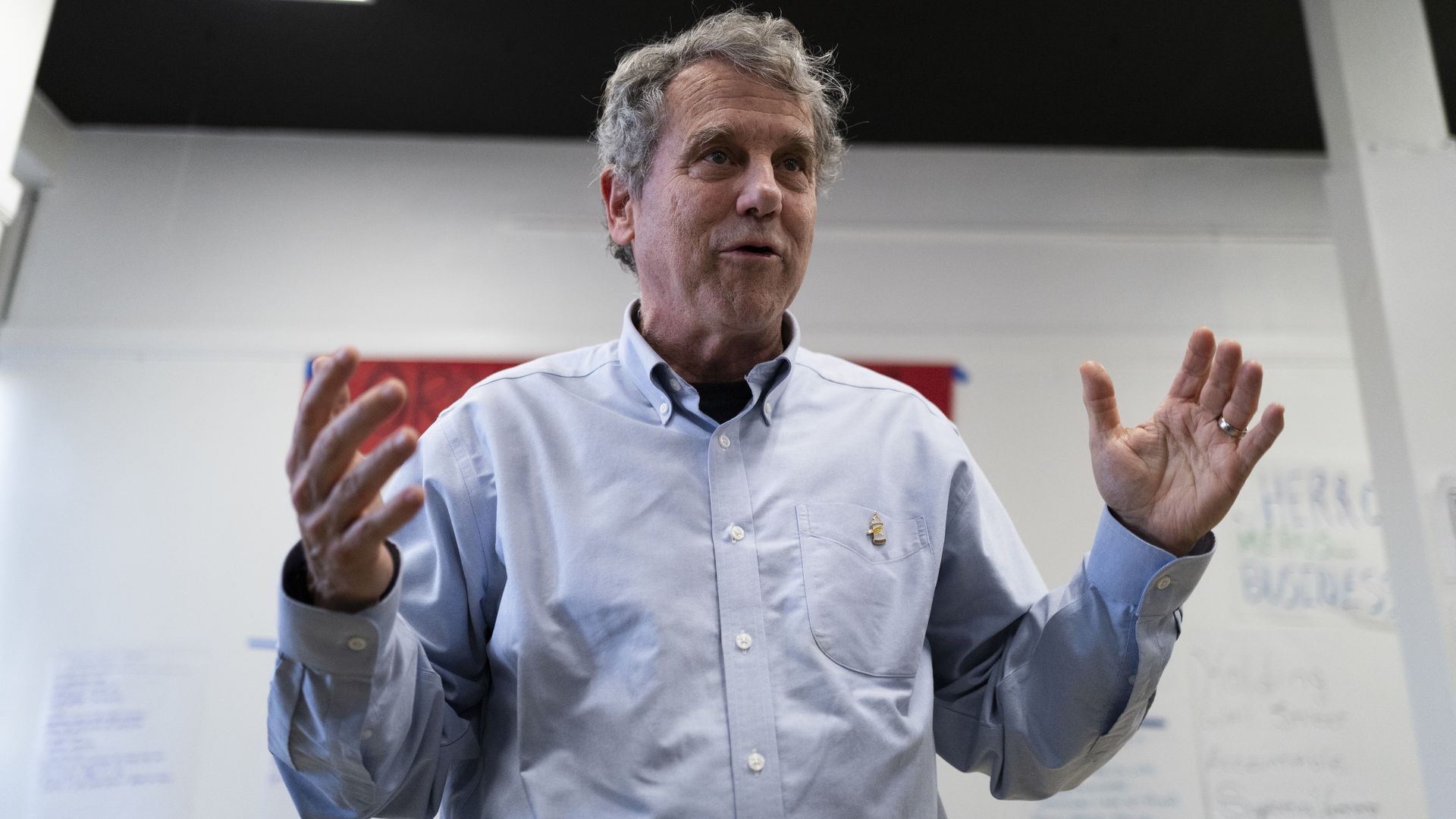 Former Sen. Sherrod Brown (D-Ohio) wearing a light blue button-up shirt speaking indoors with hands raised, against a background of white walls with text and a red horizontal stripe.
