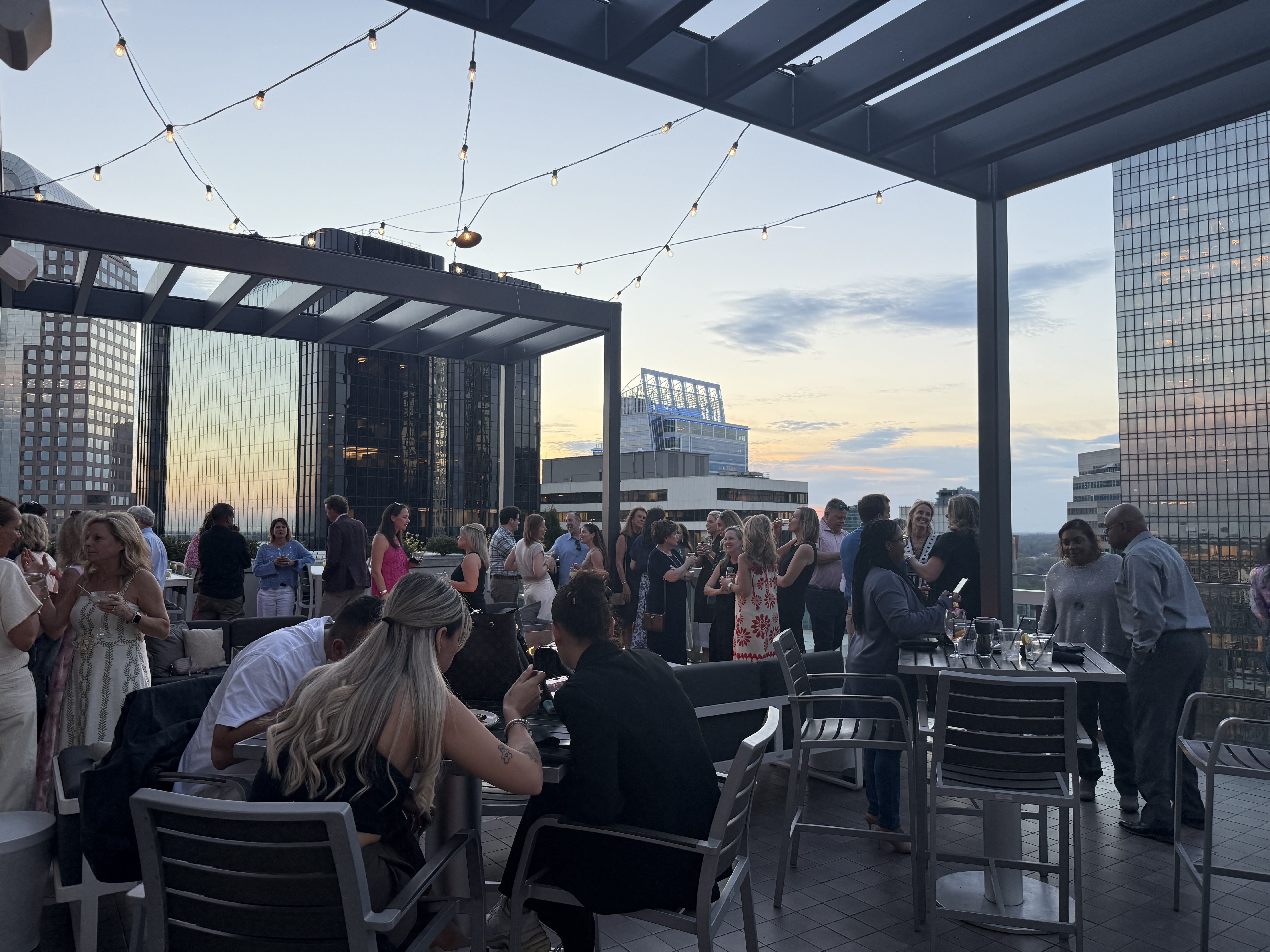 Crowded rooftop party at dusk on a city terrace. String lights crisscross overhead as guests mingle around tables and couches with tall glass office buildings in the background.