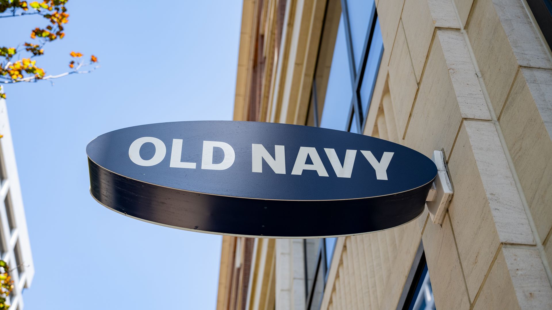 Blue oval sign reading "OLD NAVY" hangs from a beige stone building against a clear blue sky, with orange autumn leaves on a branch visible in the left foreground.